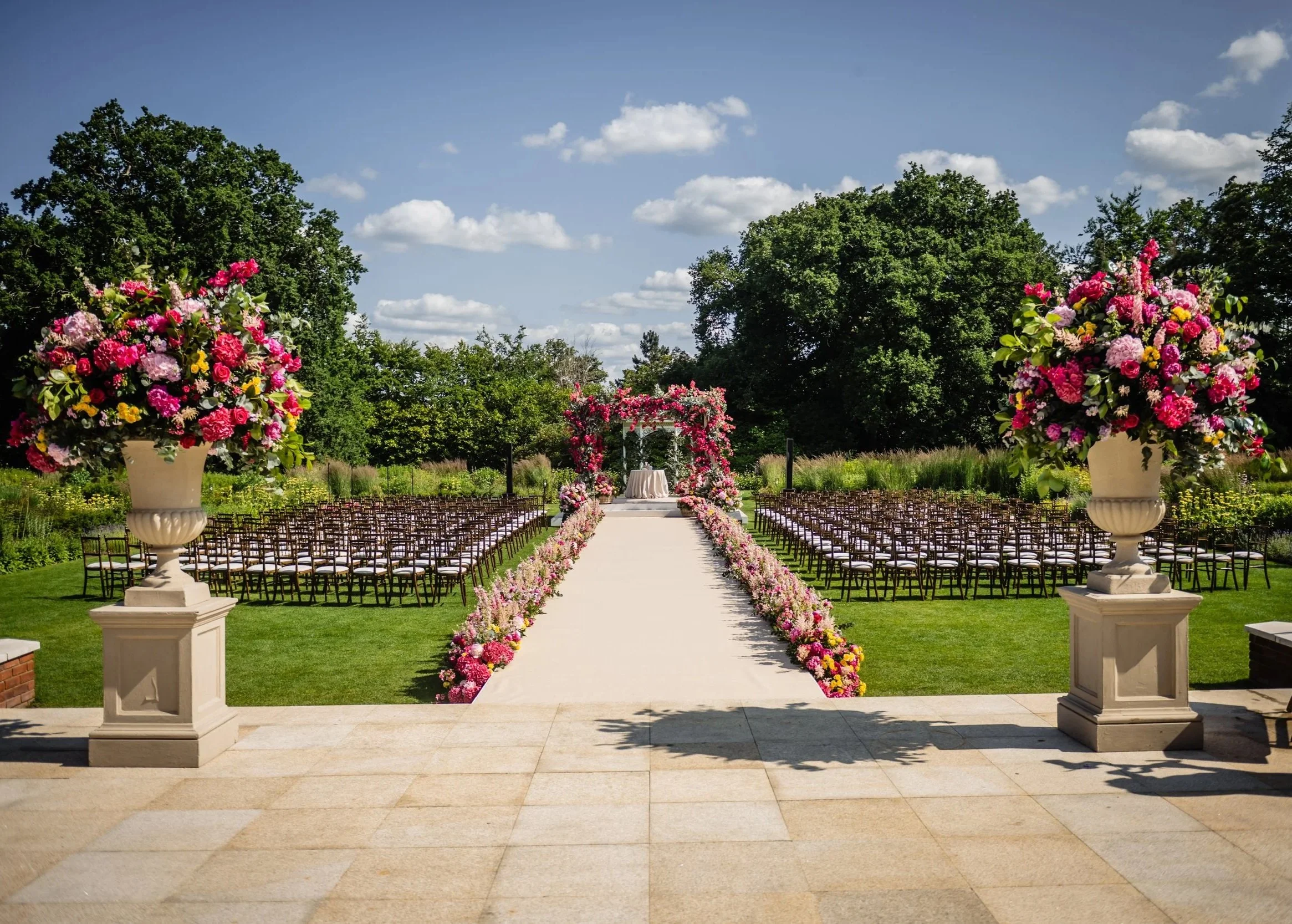 Outdoor wedding ceremony setup with a white aisle runner, floral arches, and rows of chairs in a lush green garden surrounded by trees, under a partly cloudy sky. Fairmont Windsor Park