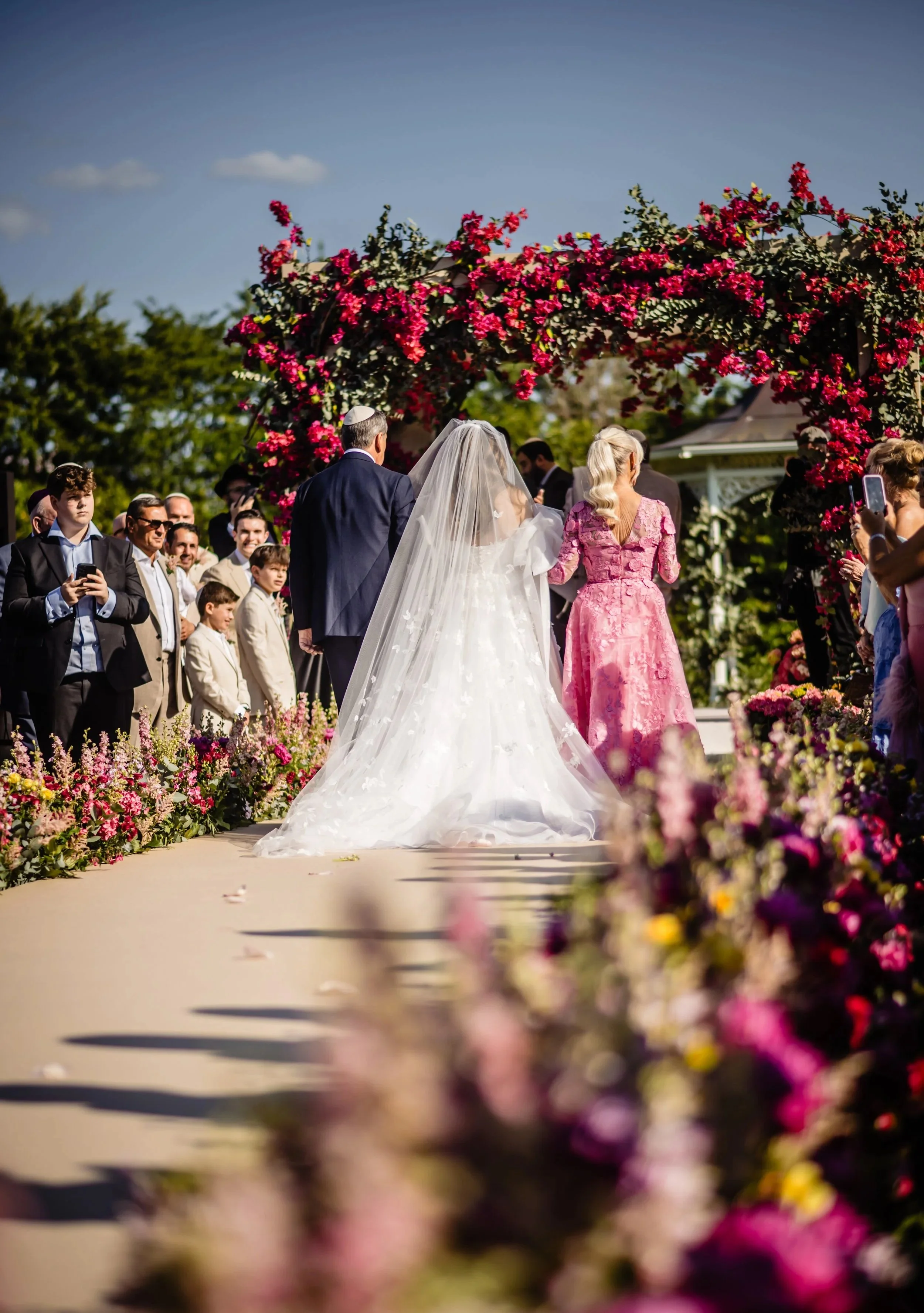A wedding ceremony taking place outdoors under a floral arch with pink flowers. The bride is in a white wedding gown and veil, standing next to a man in a dark suit, possibly the groom or a family member. Guests are gathered around, some taking photo