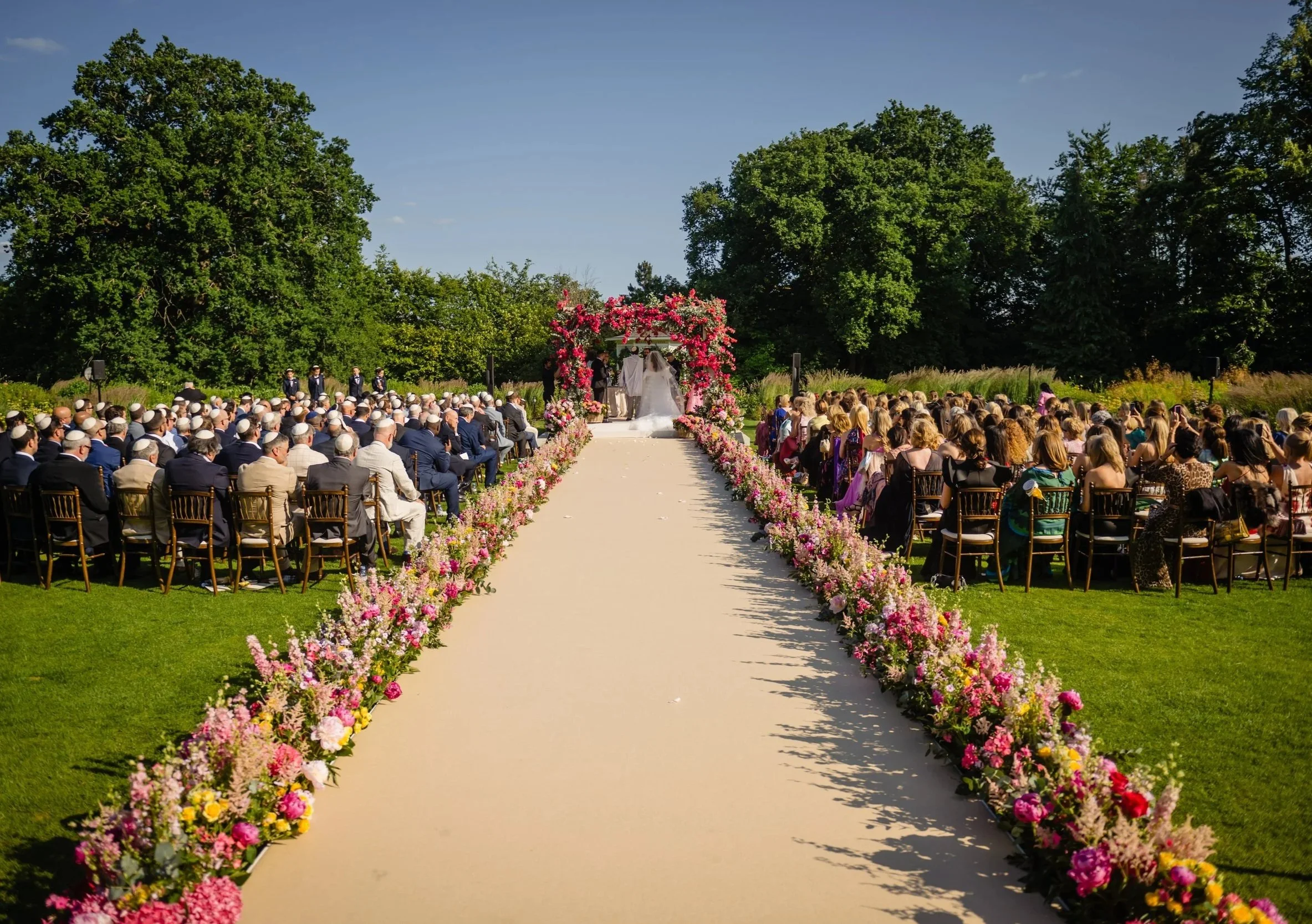 Outdoor wedding ceremony on a sunny day with guests seated on either side of a flower-adorned aisle leading to an altar where the bride and groom are exchanging vows, surrounded by green trees. Fairmont Windsor Park