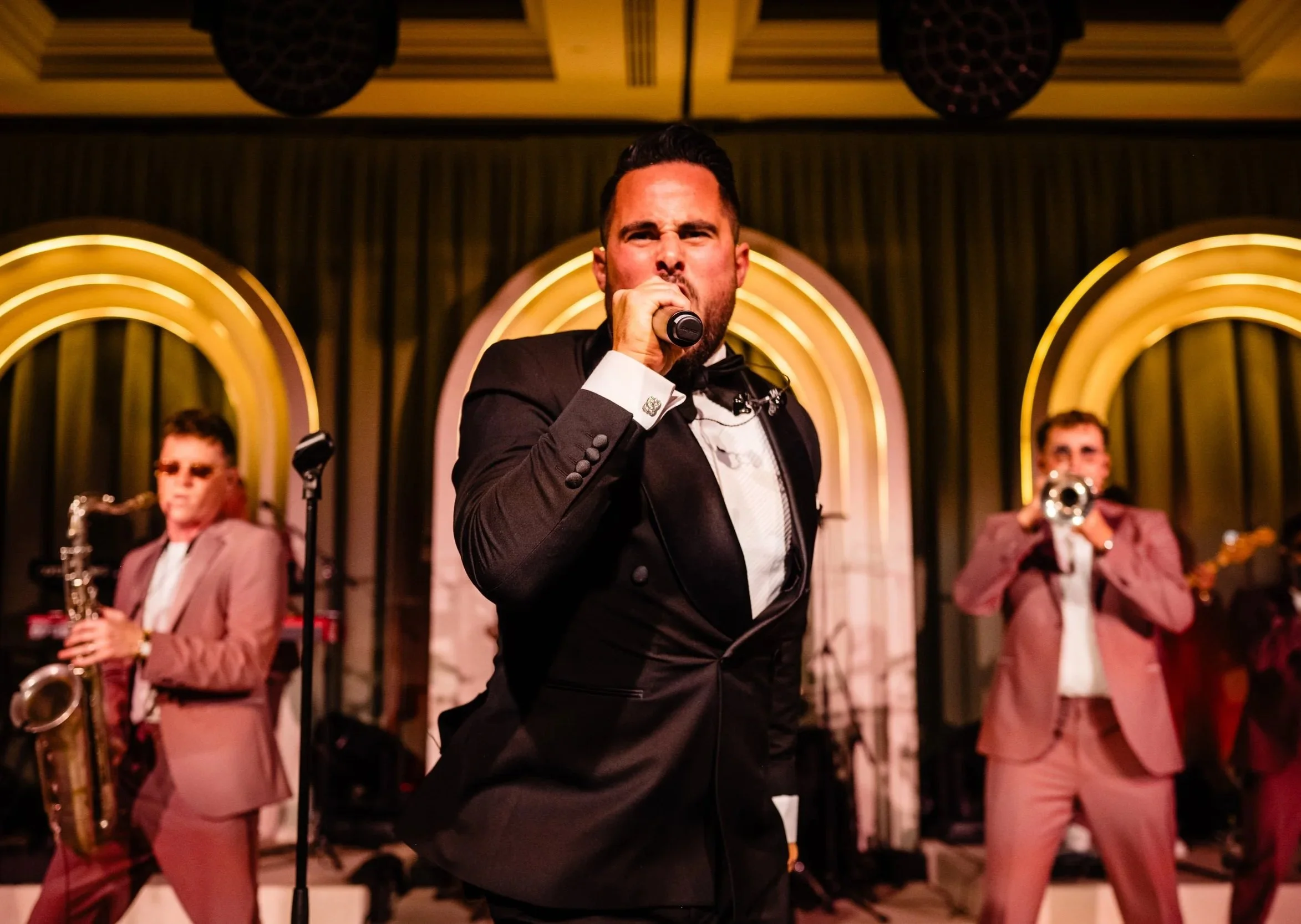 A man in a tuxedo singing into a microphone on stage, with three musicians in pink suits playing instruments behind him Fairmont Windsor Park
