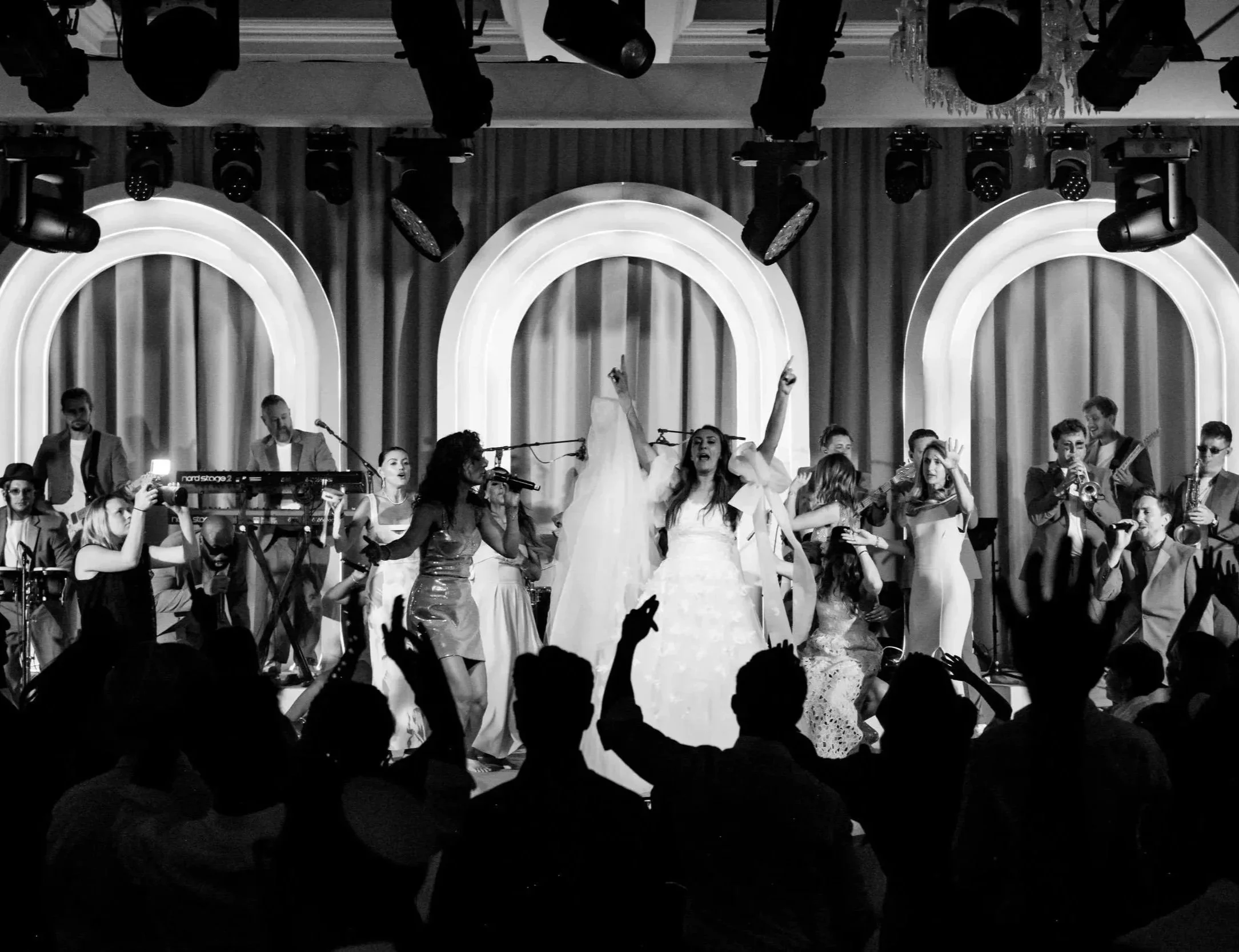 A lively stage performance with a group of singers, musicians, and dancers. Audience members are visible in the foreground, raising their hands and cheering. The scene includes stage lighting and decorative curtains Fairmont Windsor Park
