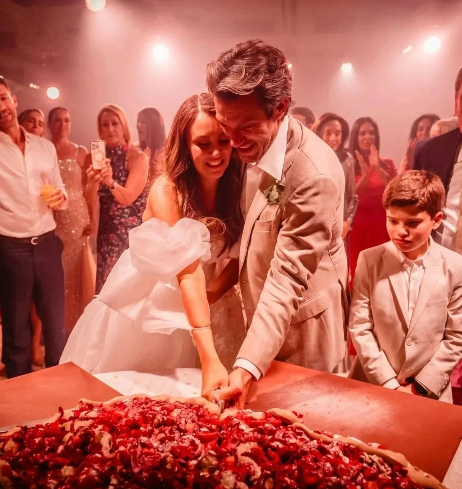 A bride and an older man are cutting a wedding cake together at a celebration. There are guests watching and smiling in the background. at Fairmont Windsor Park