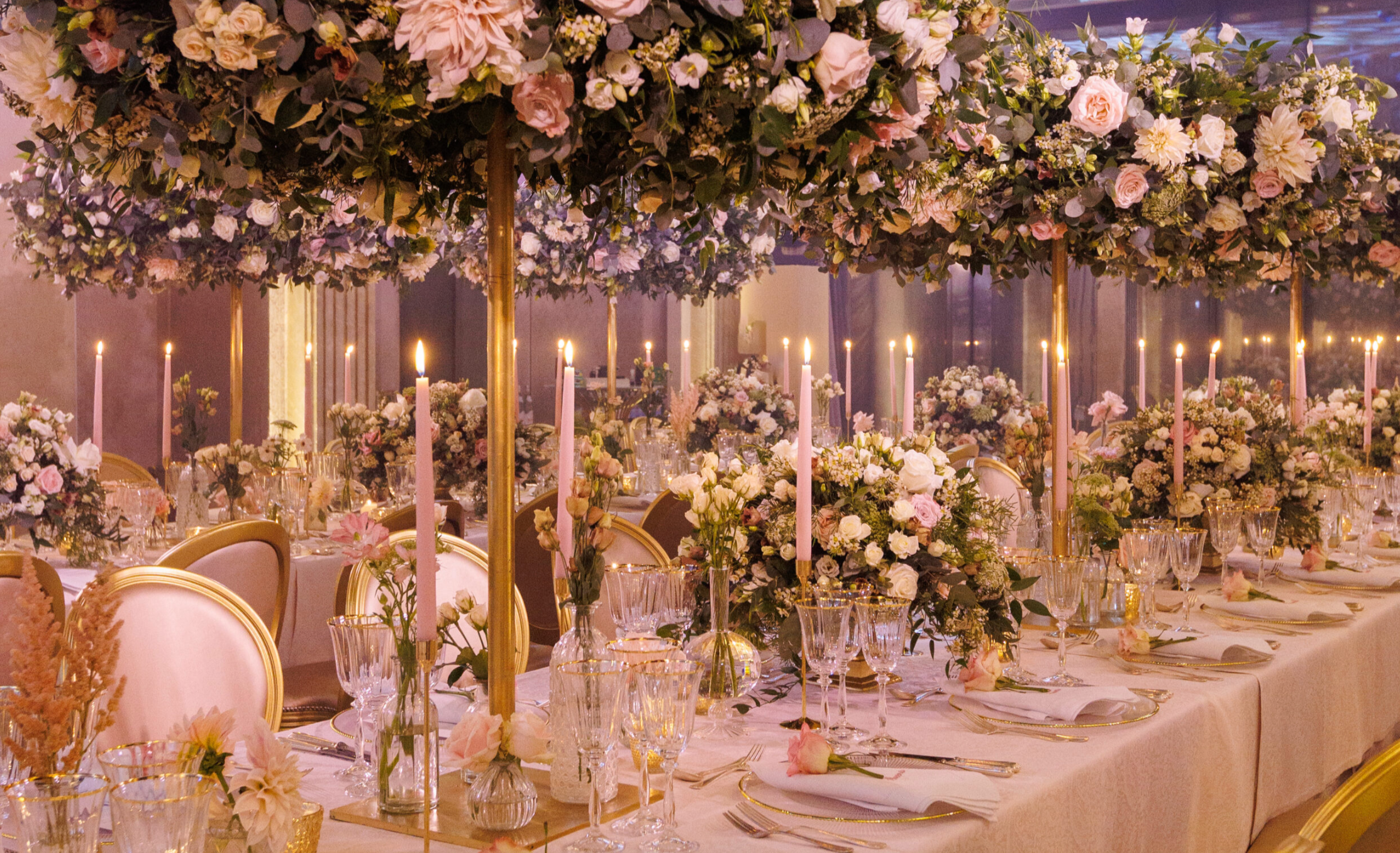 Elegant event table decorated with pink and white flowers, tall candles, and glassware, with overhead floral arrangements and warm lighting.