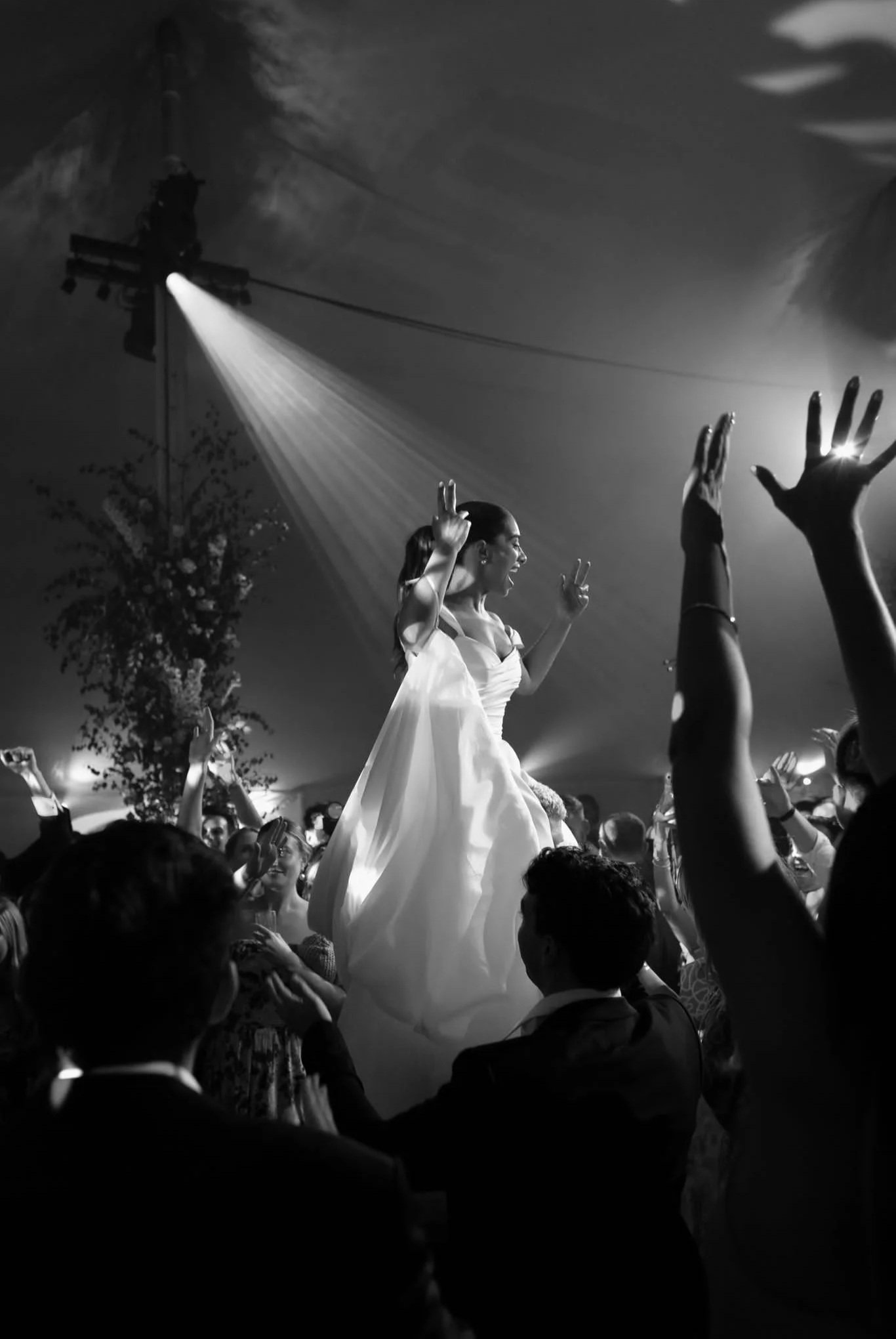 A woman in a white dress is dancing on a platform at a crowded event, with her arms raised and surrounded by people reaching out to her.