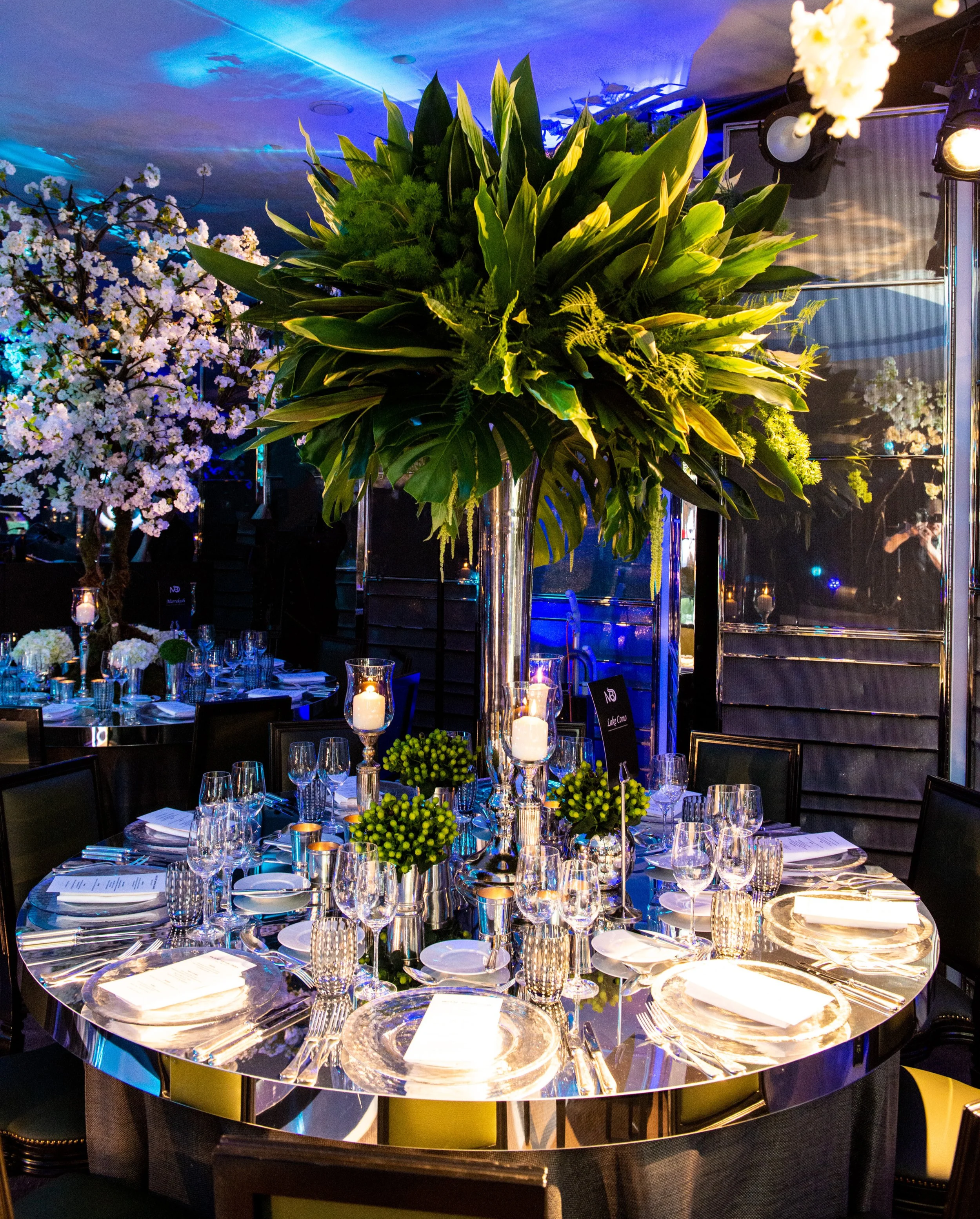 Elegant round dining table set for a formal event with crystal glassware, white plates, and black chairs. A tall clear vase with lush green foliage and a large centerpiece of greenery and tropical leaves is in the middle. The table is illuminated by 