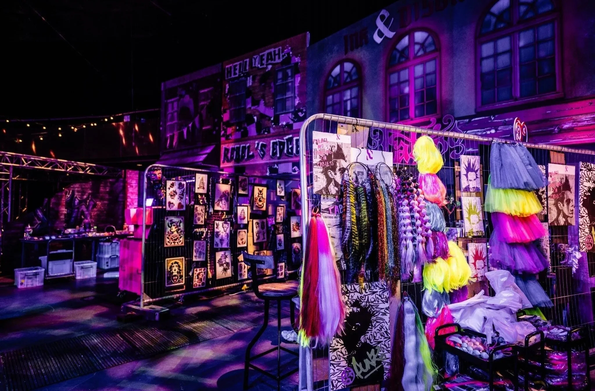A vibrant Bar Mitzvah crazy themed party selling colorful wigs, artwork, and accessories, illuminated by purple and pink lights with buildings in the background.