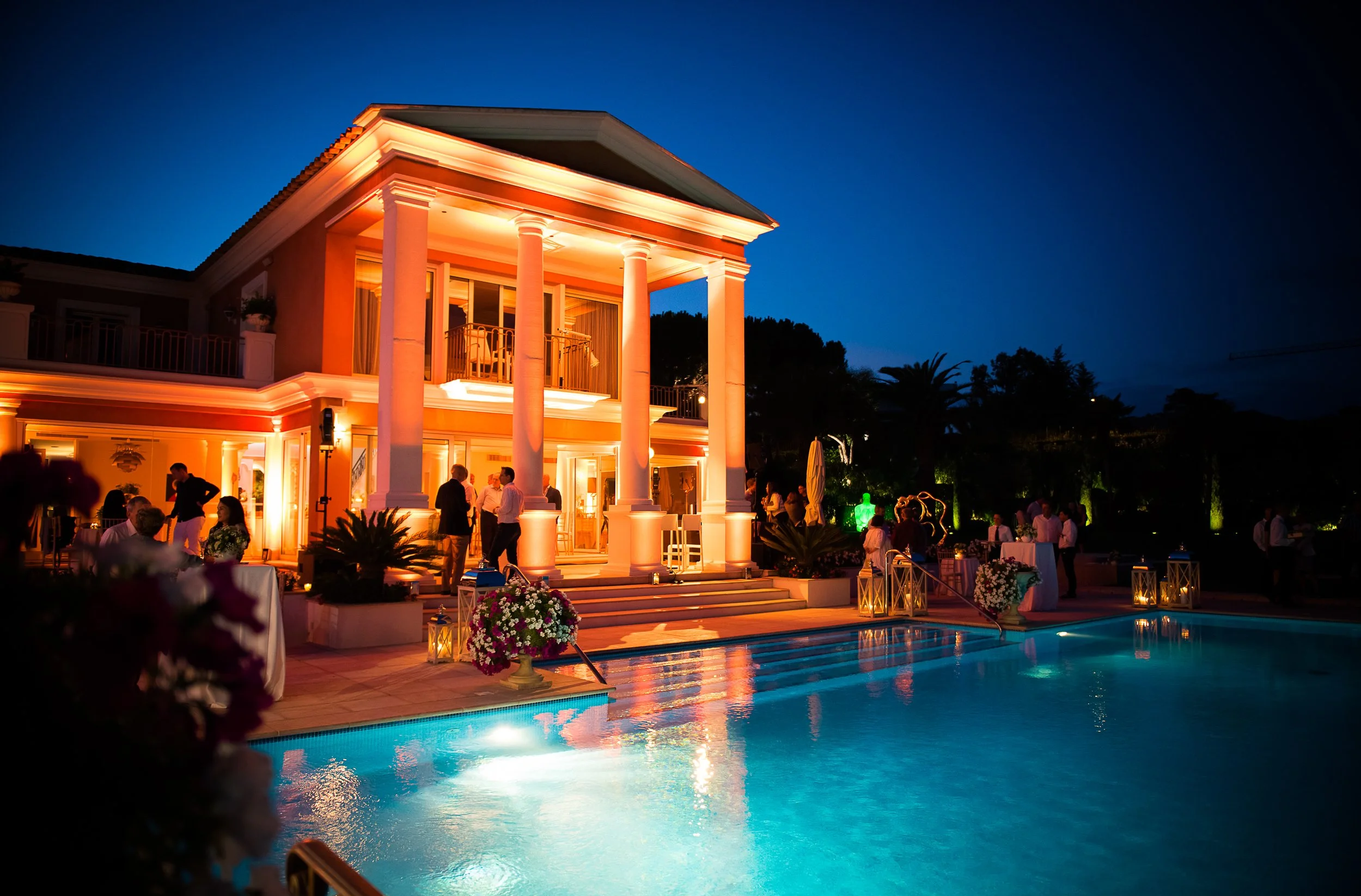 A luxurious house with white columns and a porch illuminated at night, overlooking a swimming pool with clear blue water, surrounded by greenery and poolside seating, at a social gathering.