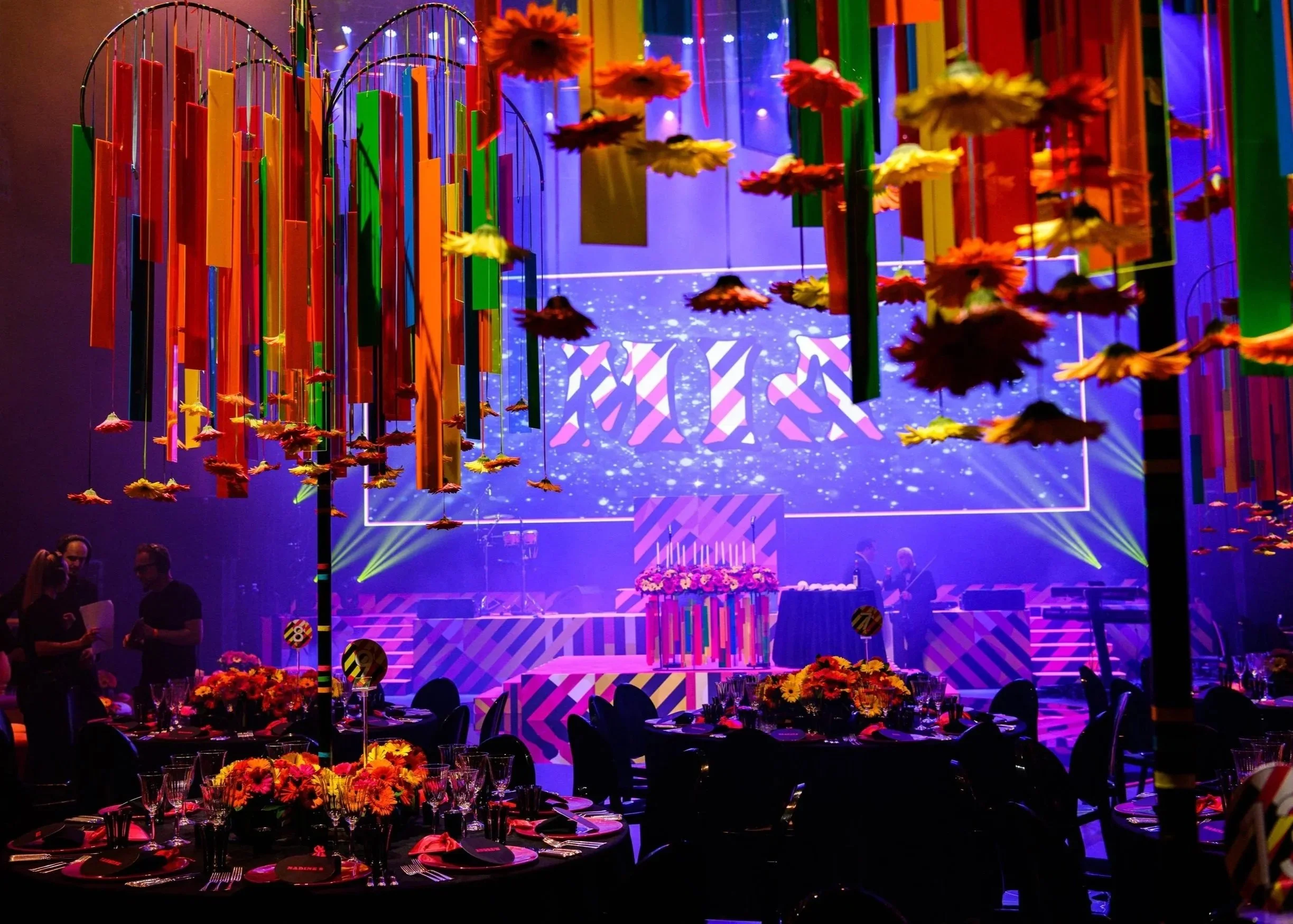 Colorful indoor event space with hanging decorations, round tables with floral centerpieces, and a stage with a large screen showing geometric patterns.  Bat Mizvah the roundhouse