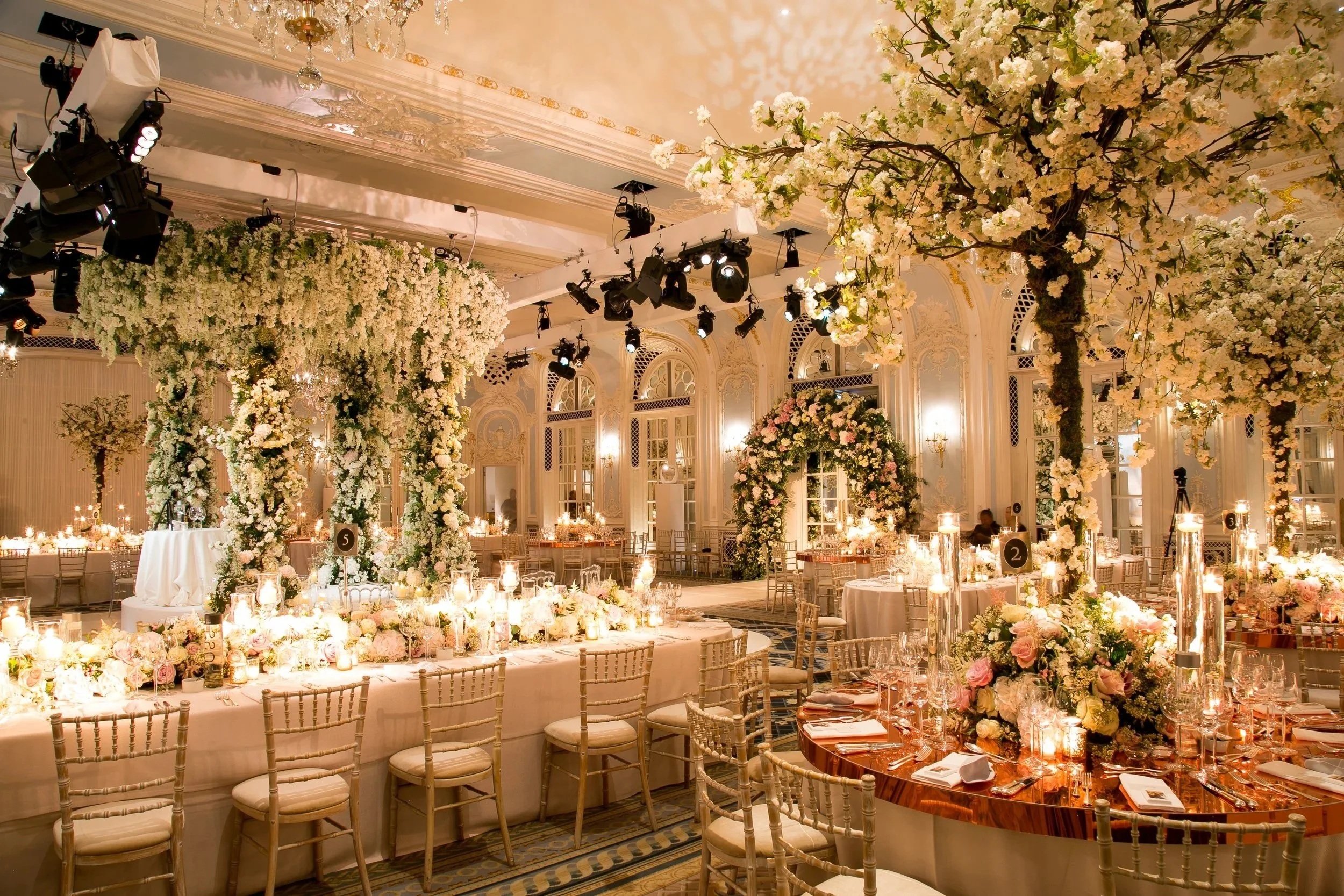 Elegant banquet hall decorated with numerous white and blush pink floral arrangements, including large flowering trees, and set with candles and fine tableware for a wedding or formal event. The ceiling has ornate moldings and a chandelier, and there