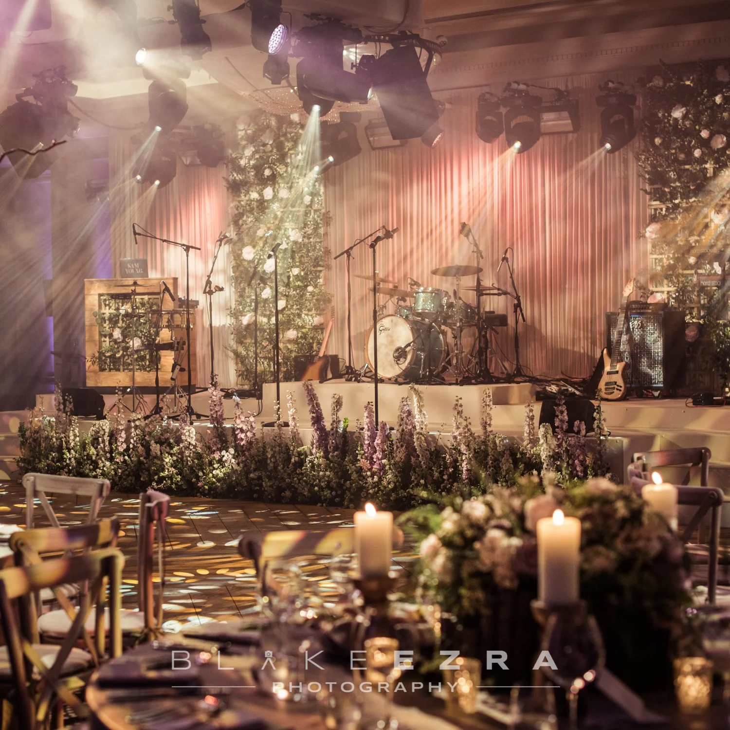 Stage with musical instruments including drums, guitars, and microphones, decorated with floral arrangements and soft lighting, set for a wedding or event.