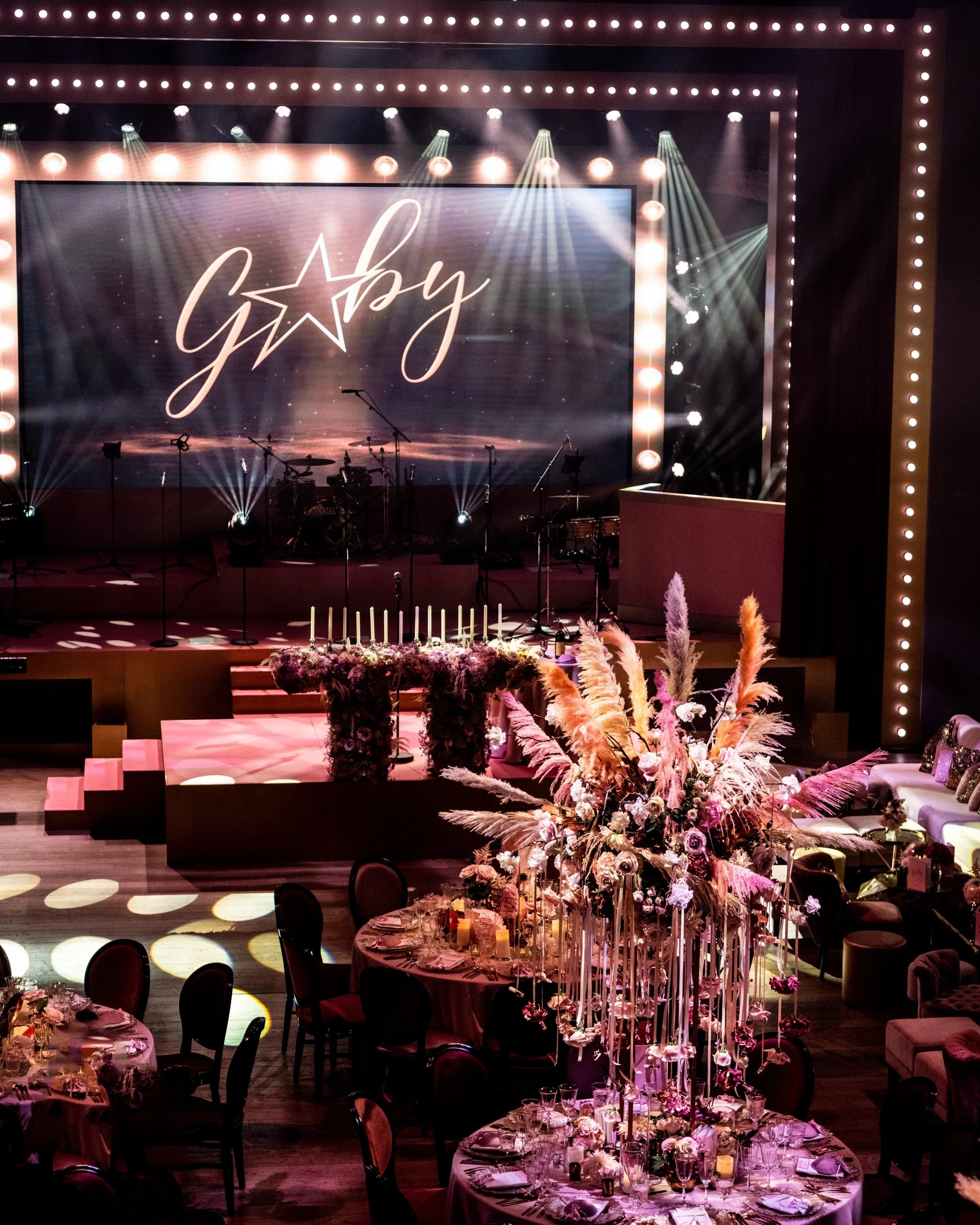 BAT MITZVAH AT BATTERSEA ARTS CENTRE. A large stage with a backdrop displaying the word 'Glamour' in cursive script, framed by bright lights and spotlights, suggesting a glamorous occasion.