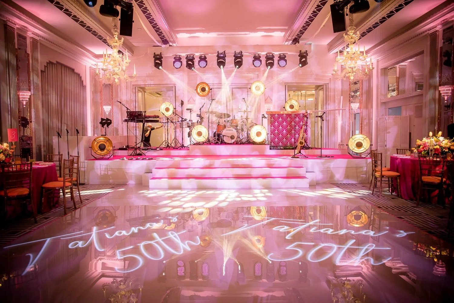 Elegant event space with a pink-lit stage set for a musical performance with drums, guitars, and microphones. The room features chandeliers, ornate wall moldings, and floral decor. The polished floor reflects pink lighting and a neon sign reading 'Ta