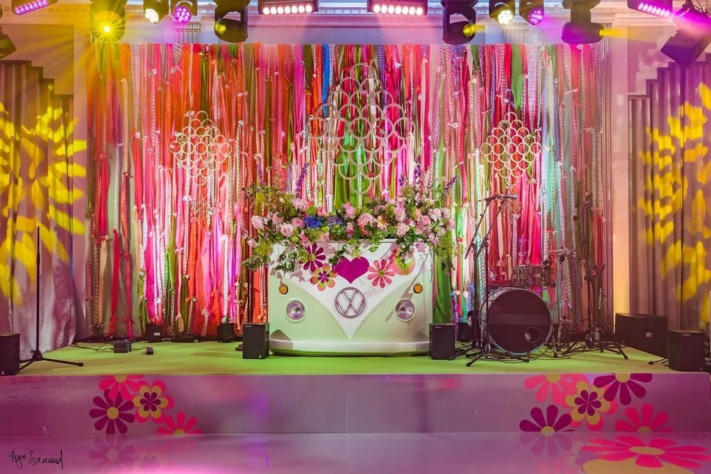 Stage decorated with colorful ribbons and flowers, featuring a floral arrangement on a white DJ booth with peace and flower symbols, surrounded by musical instruments and microphones, illuminated by colorful stage lights.