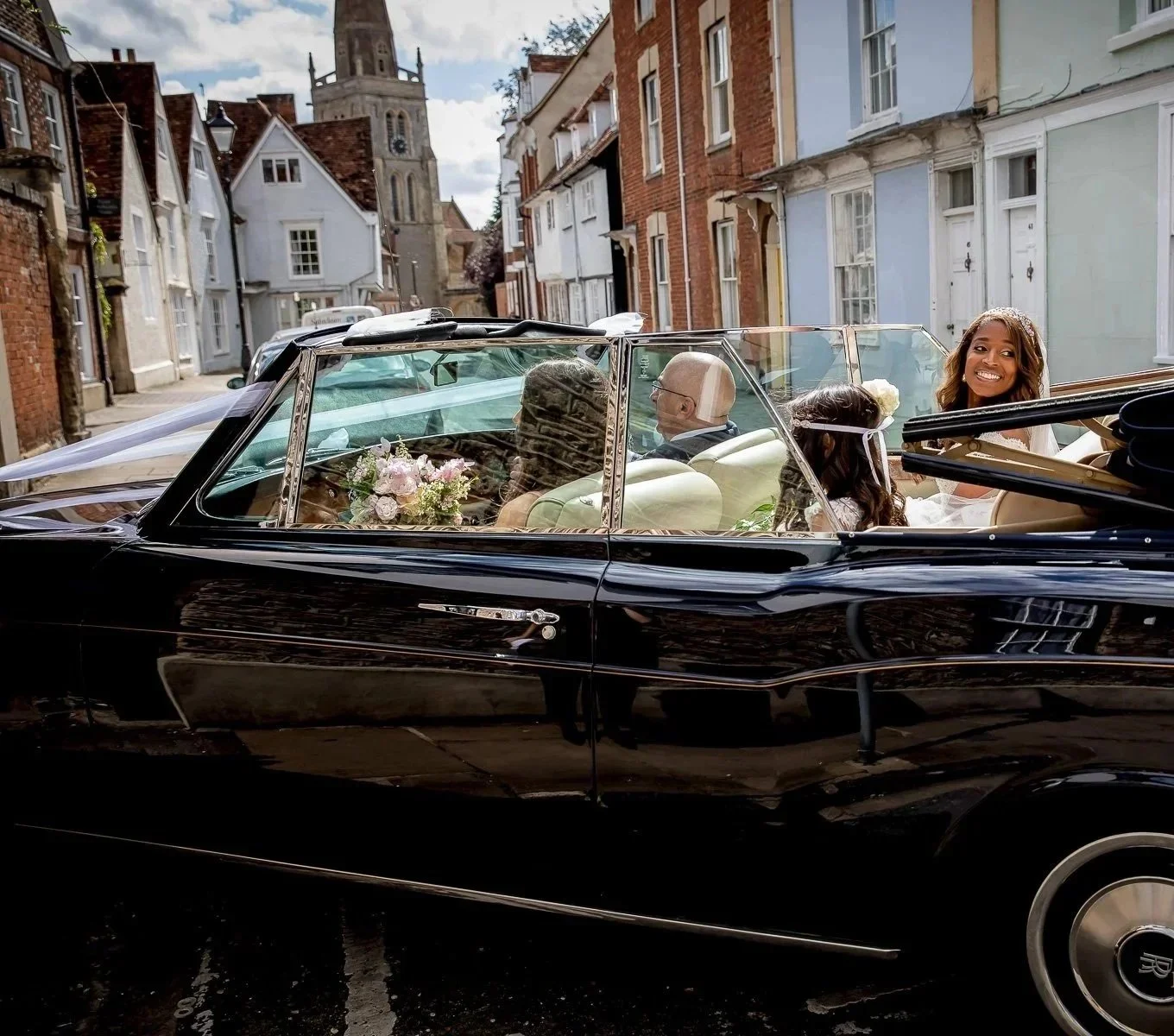 A black vintage convertible car with four passengers, including a woman with curly hair smiling at the camera, driving through a cobblestone street with colorful old buildings and a church in the background. The car is decorated with flowers, suggest