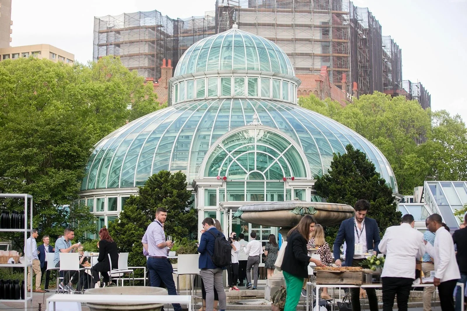 Group of people gathered outdoors in front of a large glass conservatory with a domed roof, surrounded by trees, with high-rise buildings in the background during daytime.