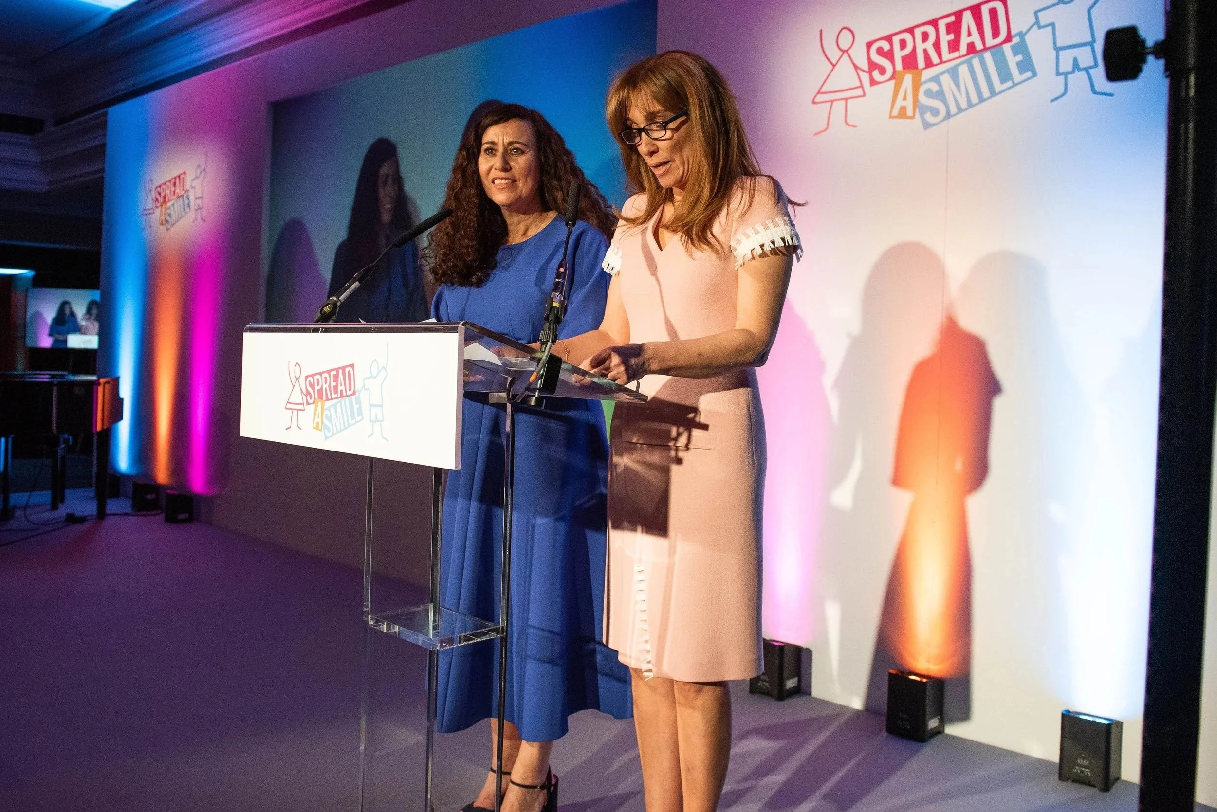 Two women standing at a podium with a microphone on a stage at an event with a large backdrop. The backdrop has a logo and the slogan 'Spread a Smile' along with colorful illustrations of smiling figures. One woman is wearing a blue dress, and the ot
