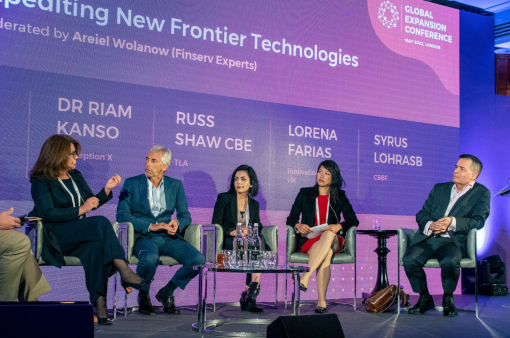 Panel discussion at a conference with five speakers seated on stage. The backdrop features a large screen displaying names and titles, and the conference is the Global Expansion Conference, held in London, May 2022.