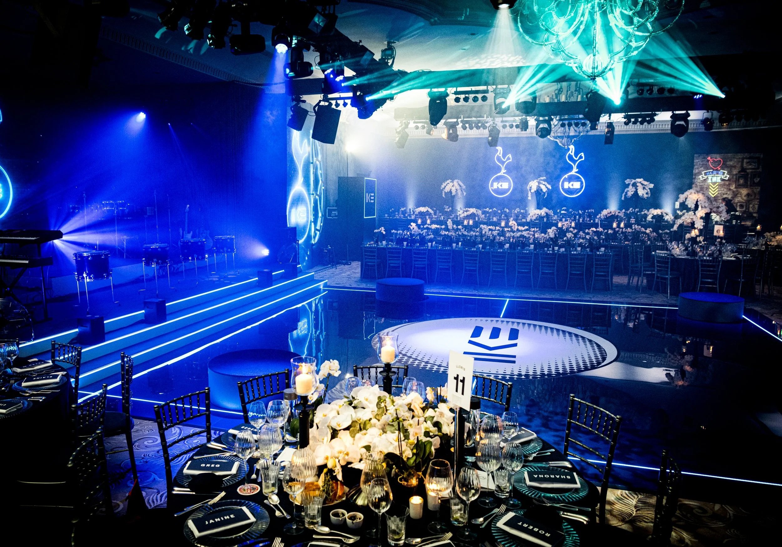 A decorated event venue with a stage illuminated by blue and white lights, circular chandeliers, and large screens displaying logos. In the foreground, a round table is set with wine glasses, plates, silverware, napkins labeled with guest names, and 