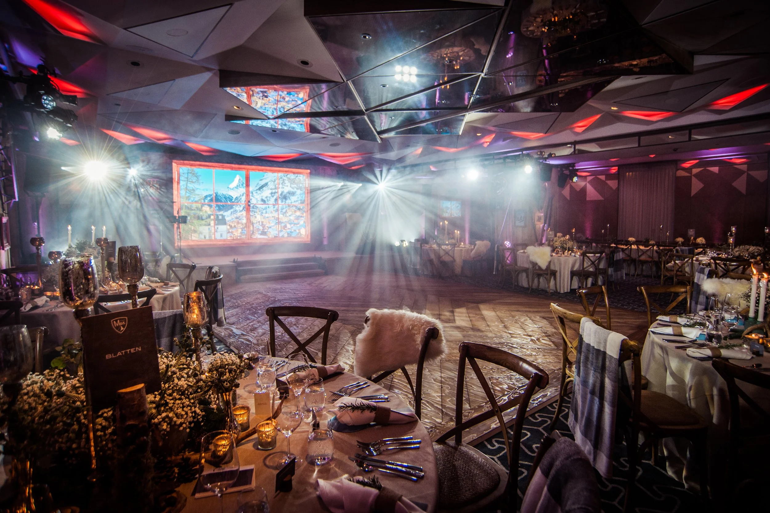 An elegantly decorated banquet hall with tables set for a formal event, featuring floral centerpieces, candles, and glassware. There is a large projection screen showing a scenic mountain landscape, and the ceiling has reflective geometric panels wit