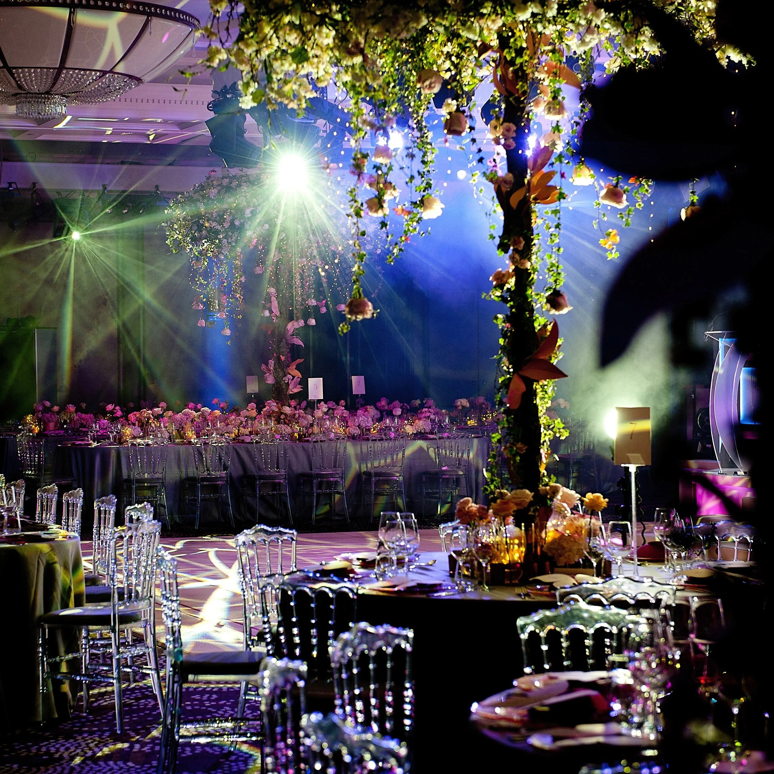 Decorated event venue with floral arrangements hanging from the ceiling, illuminated by green and purple lighting, set with tables and transparent chairs.