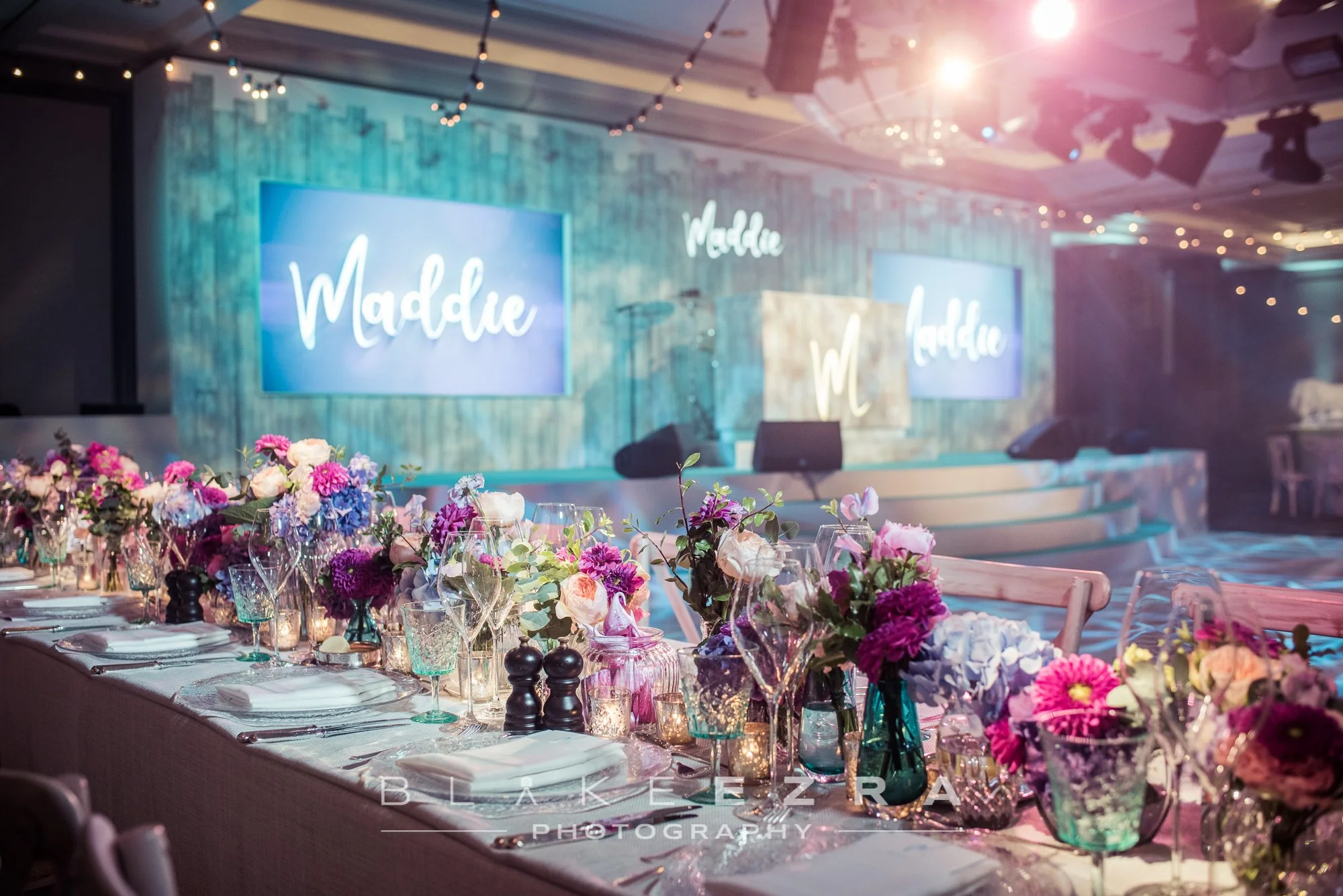 Decorated event space with a long table set with white tablecloth, glassware, plates, and floral centerpieces in purple, pink, and white, with candles. In the background, a stage with two large screens displaying the name 'Maddie' in cursive, hanging
