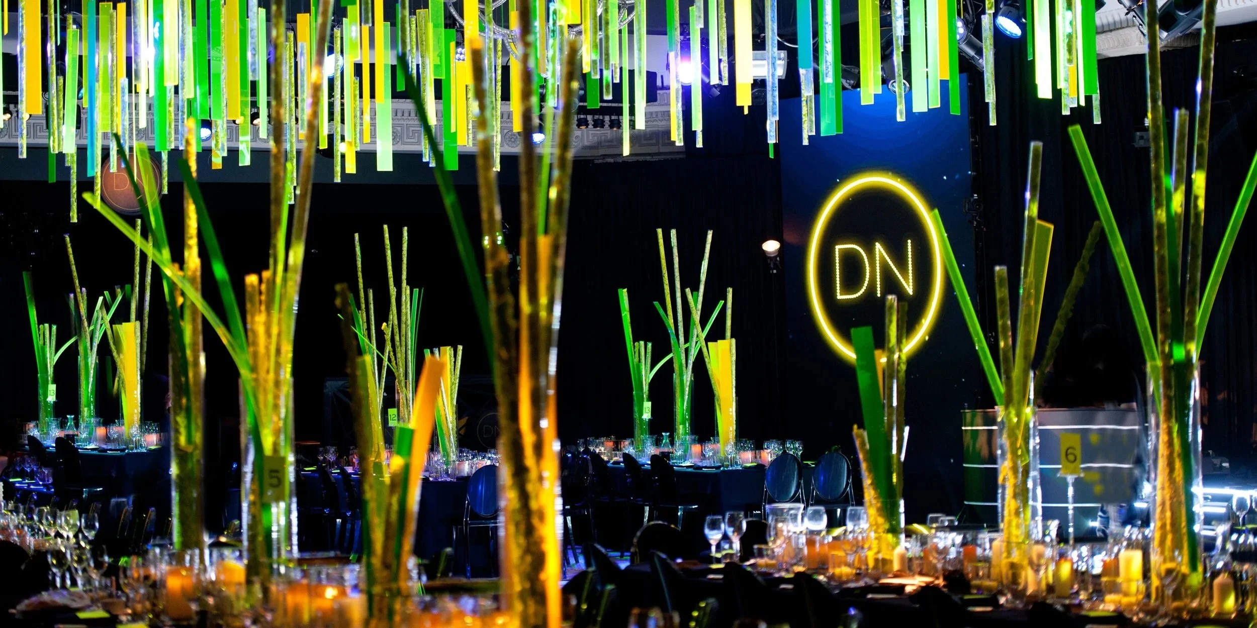A decorated event space with tall, glowing yellow and green ornamental structures, a large illuminated yellow oval sign with 'DN' on it, and tables set with glassware and candles.