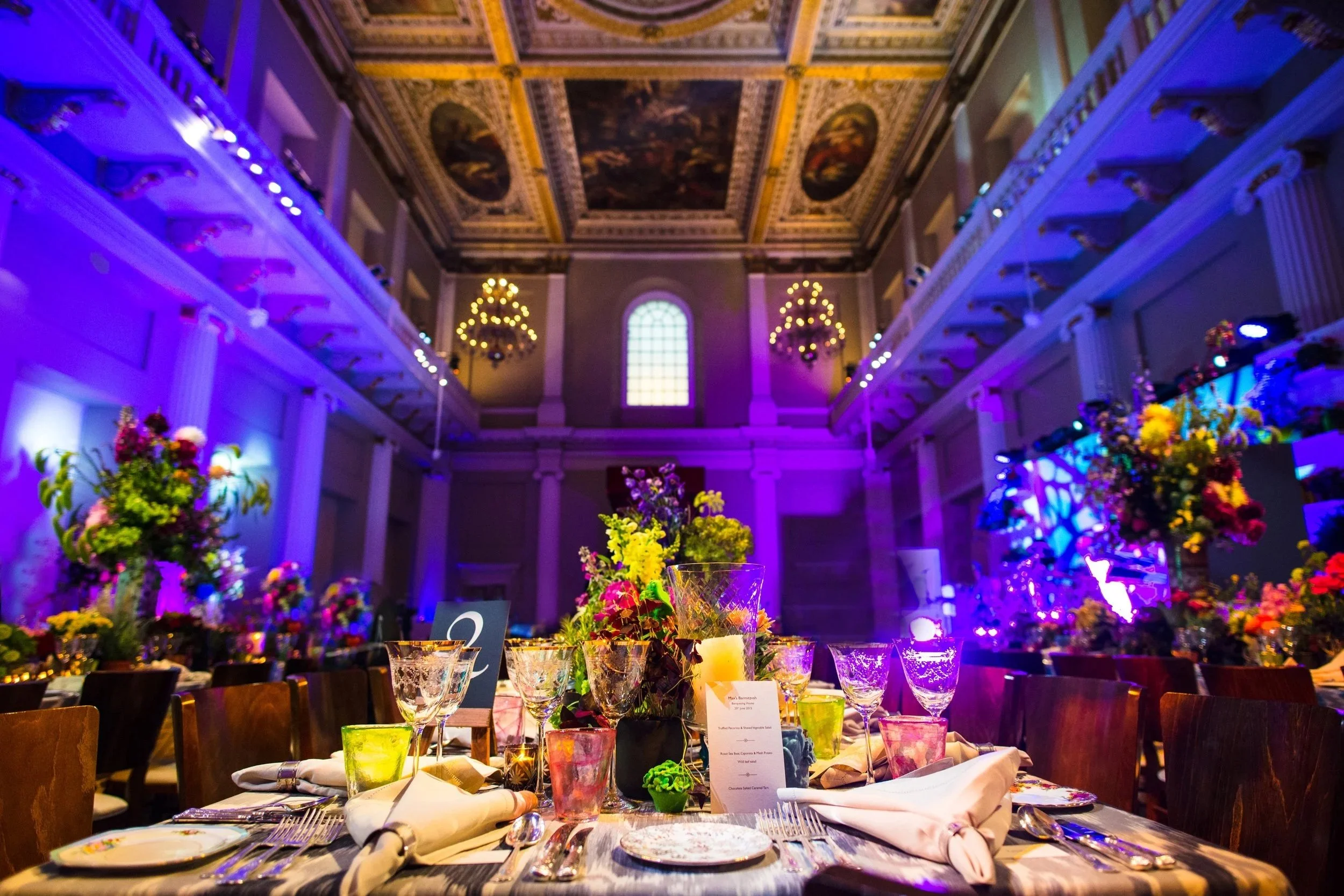 Elegant banquet hall decorated with flowers and colorful glassware on a table set for a formal event under a high, decorated ceiling with chandeliers and ample windows.