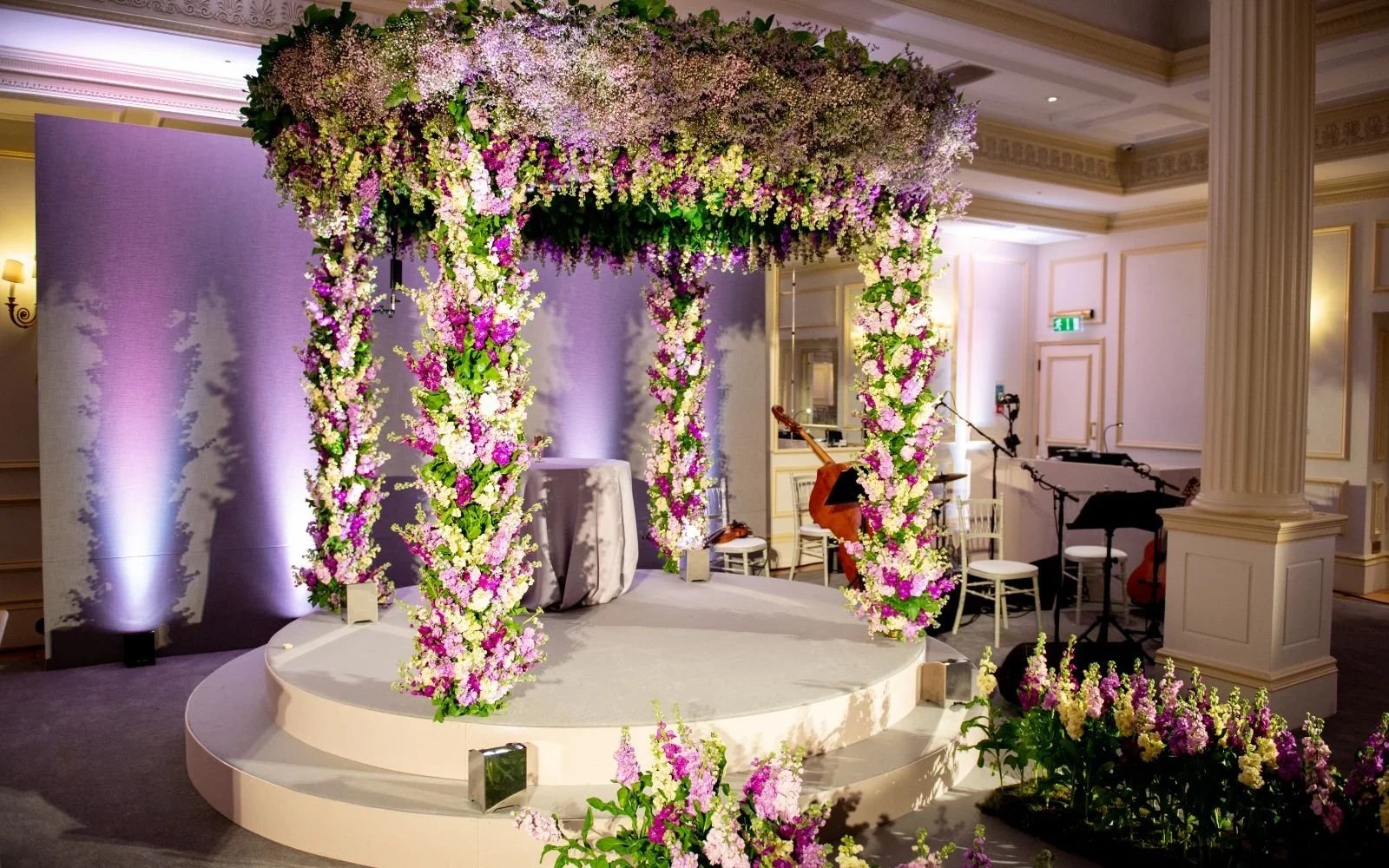A decorated stage with a floral arch made of pink, white, and purple flowers in a banquet hall for a wedding or event. There is a small round platform, a table draped in white cloth, and musical instruments including a cello, guitar, and microphones 