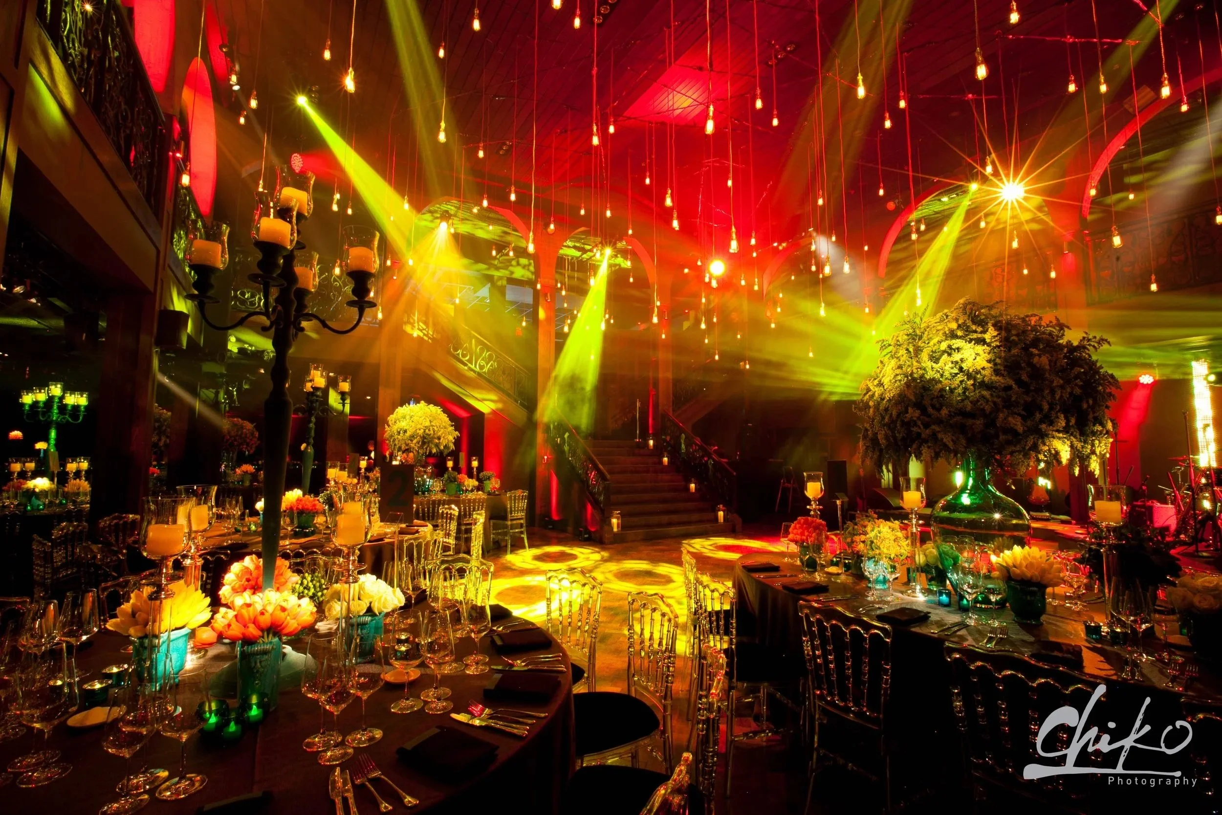 An elaborately decorated event hall with hanging lights, colorful stage lighting, floral centerpieces, and elegant table settings.