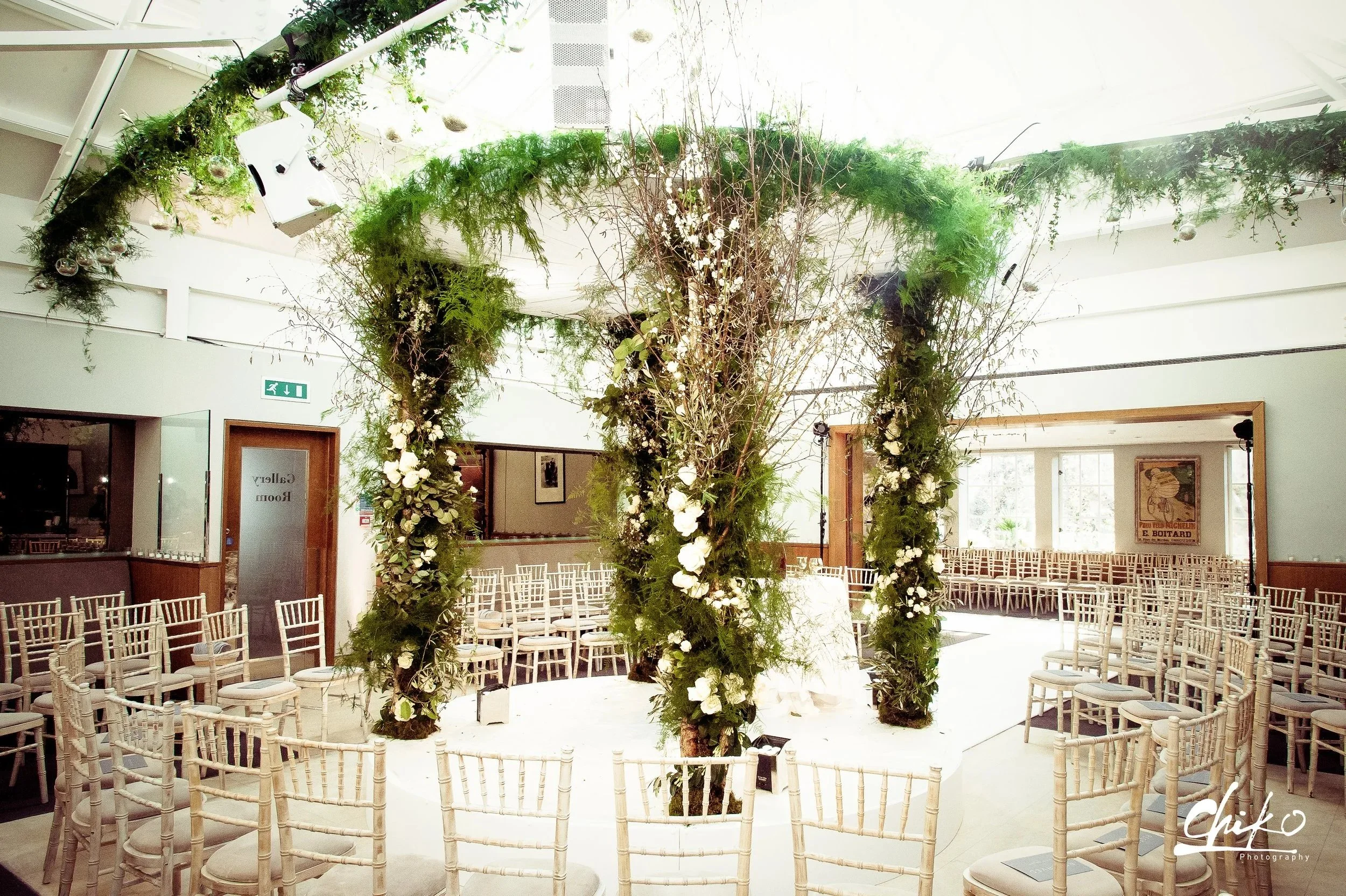 Interior of a building decorated for a wedding or event, with a floral arch and numerous white chairs arranged in rows.