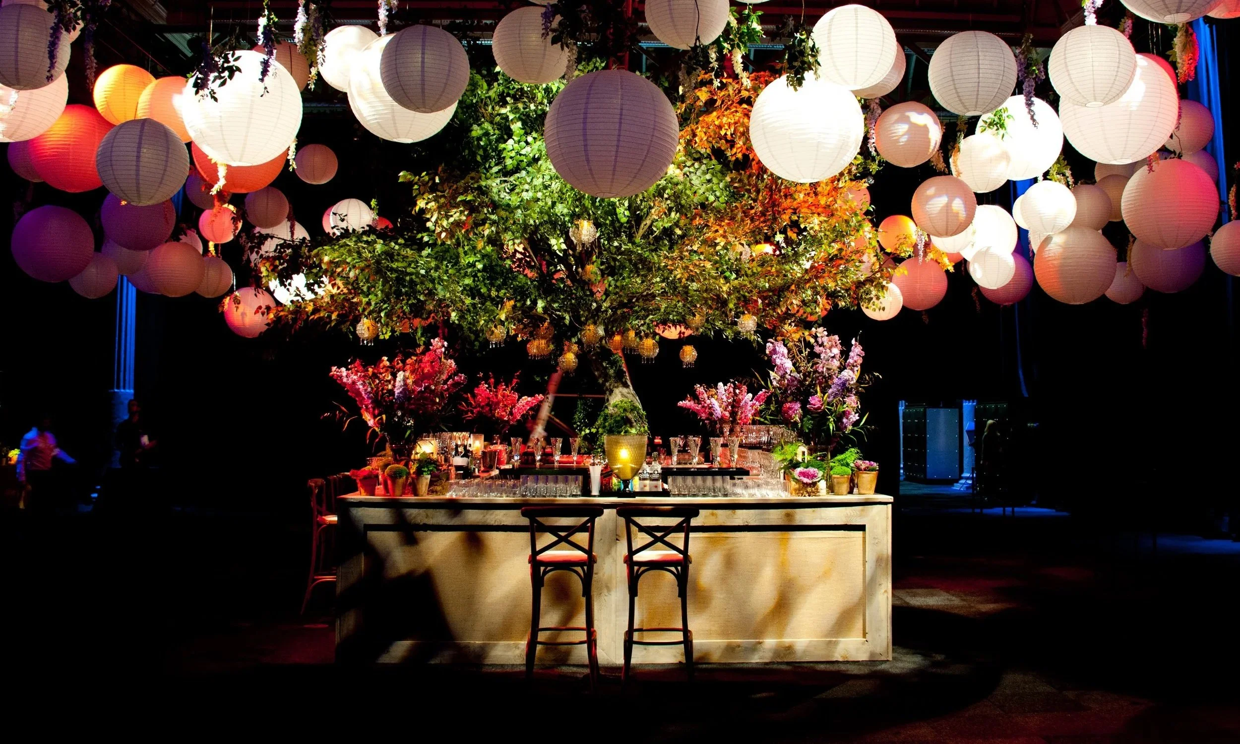 A decorated event space with hanging paper lanterns in shades of white, pink, and purple, and colorful flowers arranged on a central structure, illuminated by soft lighting.