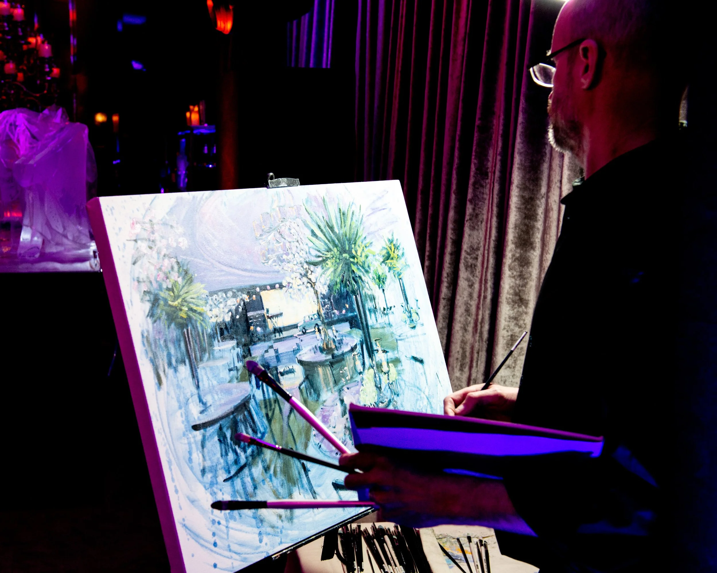 Person painting a scene with palm trees and a fountain on an easel in a dimly lit environment with purple and pink lighting.