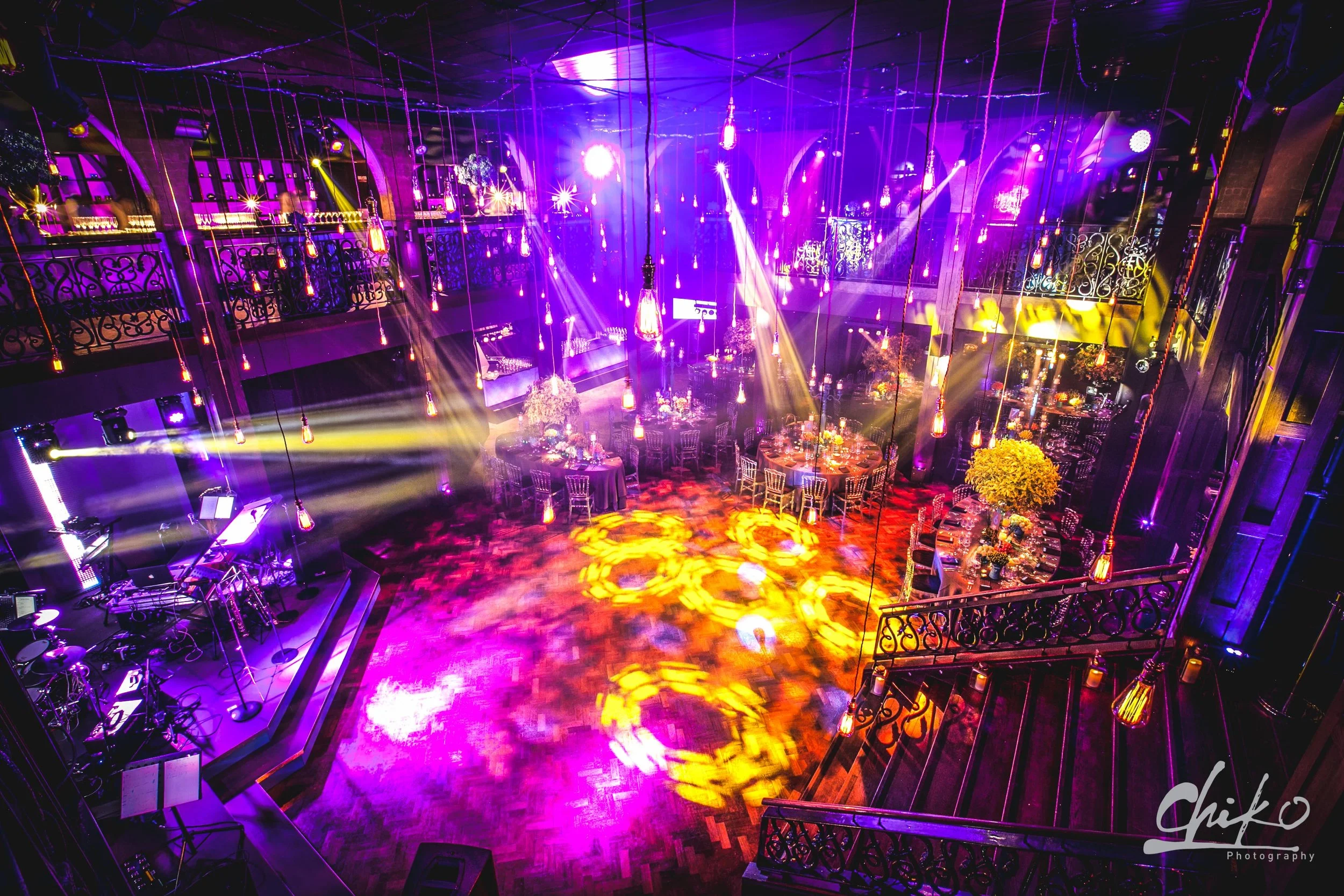 A dimly lit indoor event space with purple and yellow stage lighting, decorated tables, and hanging Edison-style light bulbs creating a festive atmosphere.