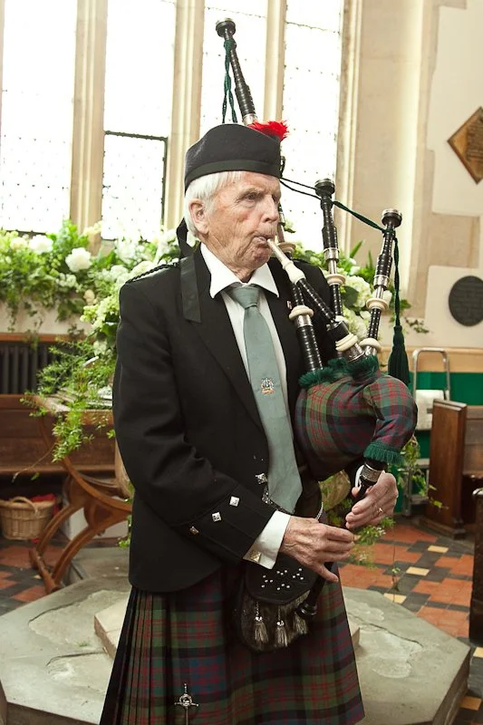 An elderly man dressed in traditional Scottish attire, playing the bagpipes in a bright, indoor setting with plants and wooden furniture.