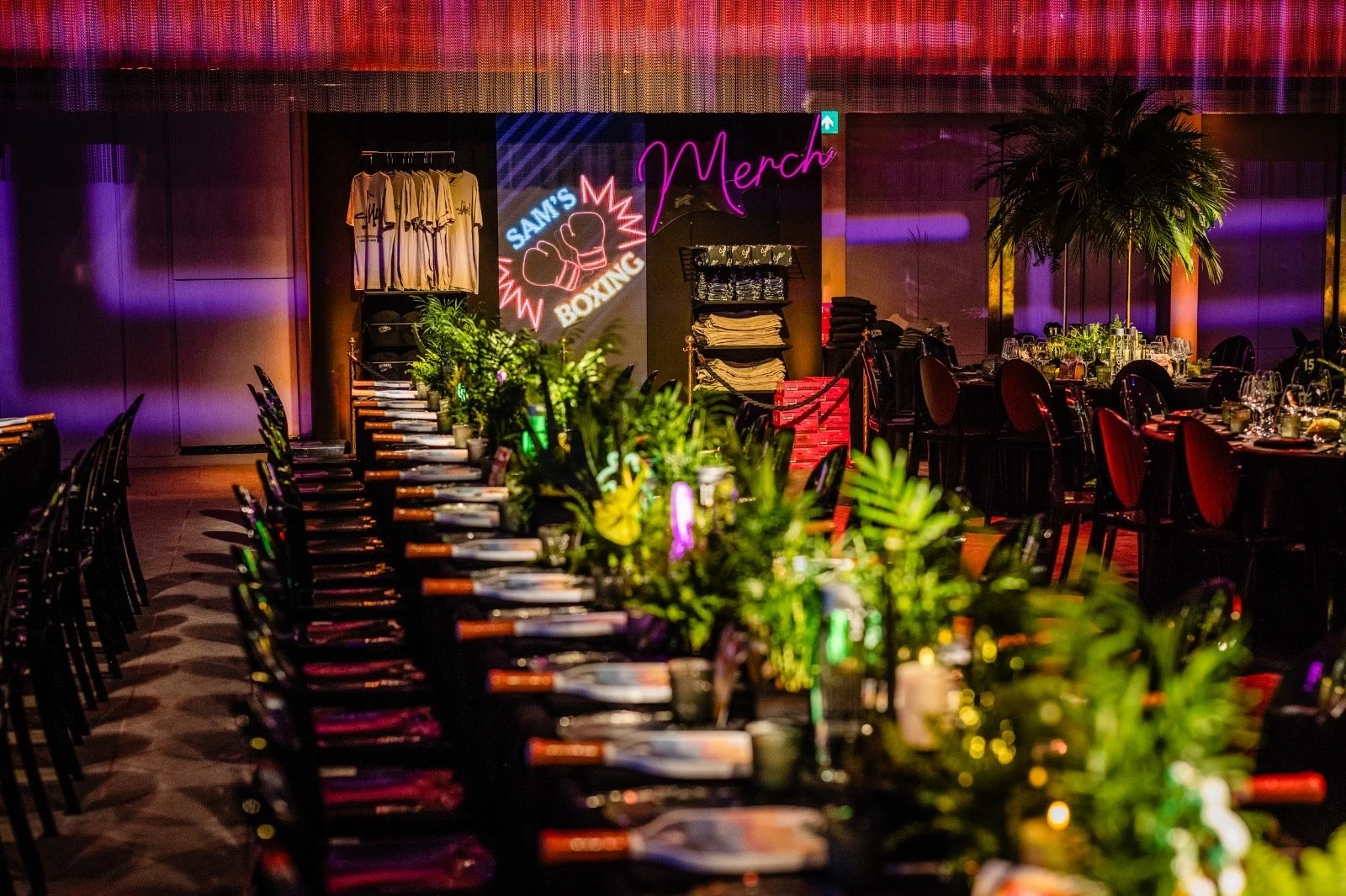A decorated indoor banquet hall with long dining tables set with plates, glasses, and cutlery, illuminated by colorful lighting, with a backdrop of neon signs and lush greenery Bar Mitzvah