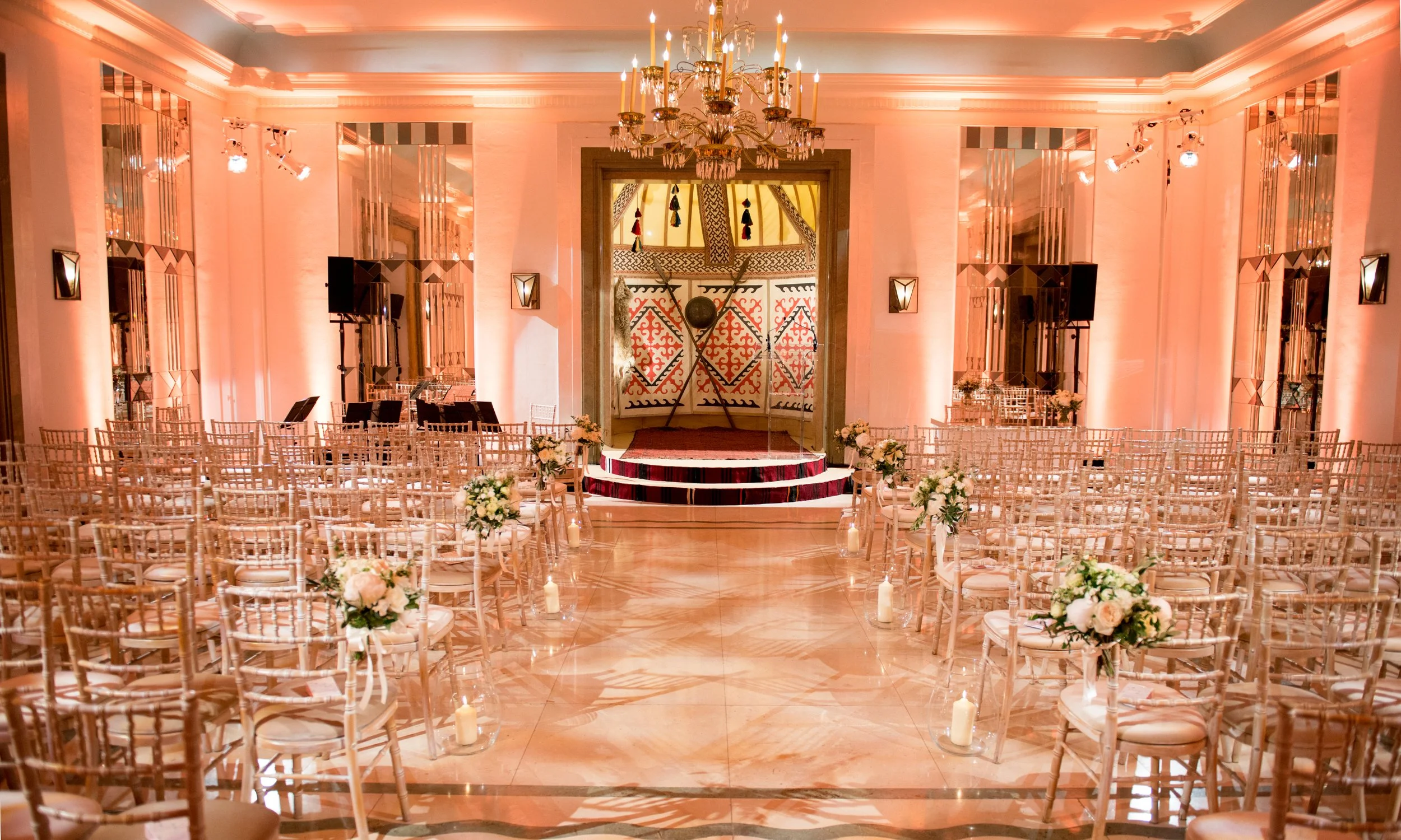 Elegant wedding ceremony setup in a luxurious ballroom with rows of white chairs decorated with flowers, a red carpeted aisle, candles along the aisle, a chandelier hanging from the ceiling, and a decorated arch at the front.