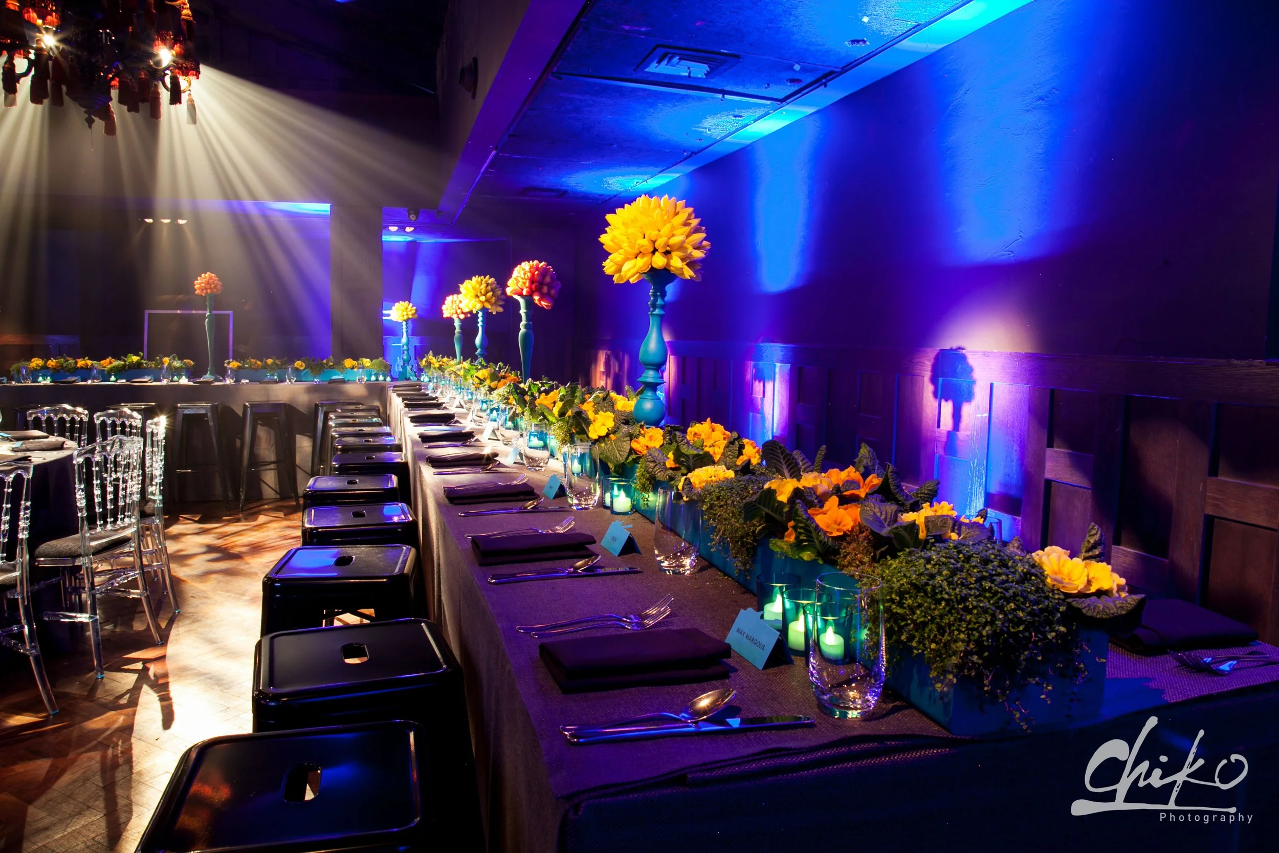 A long banquet table decorated with yellow flowers in tall vases and small candles, set for an event in a dimly lit room with blue lighting and a dark ceiling.