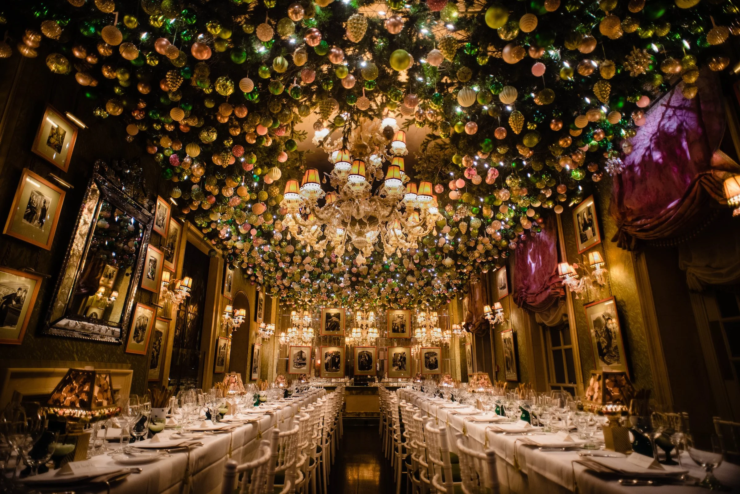 A luxurious dinner banquet hall decorated for the holidays, featuring a ceiling covered with colorful ornaments and Christmas lights, a grand chandelier, and tables set with white tablecloths, glassware, and chairs.