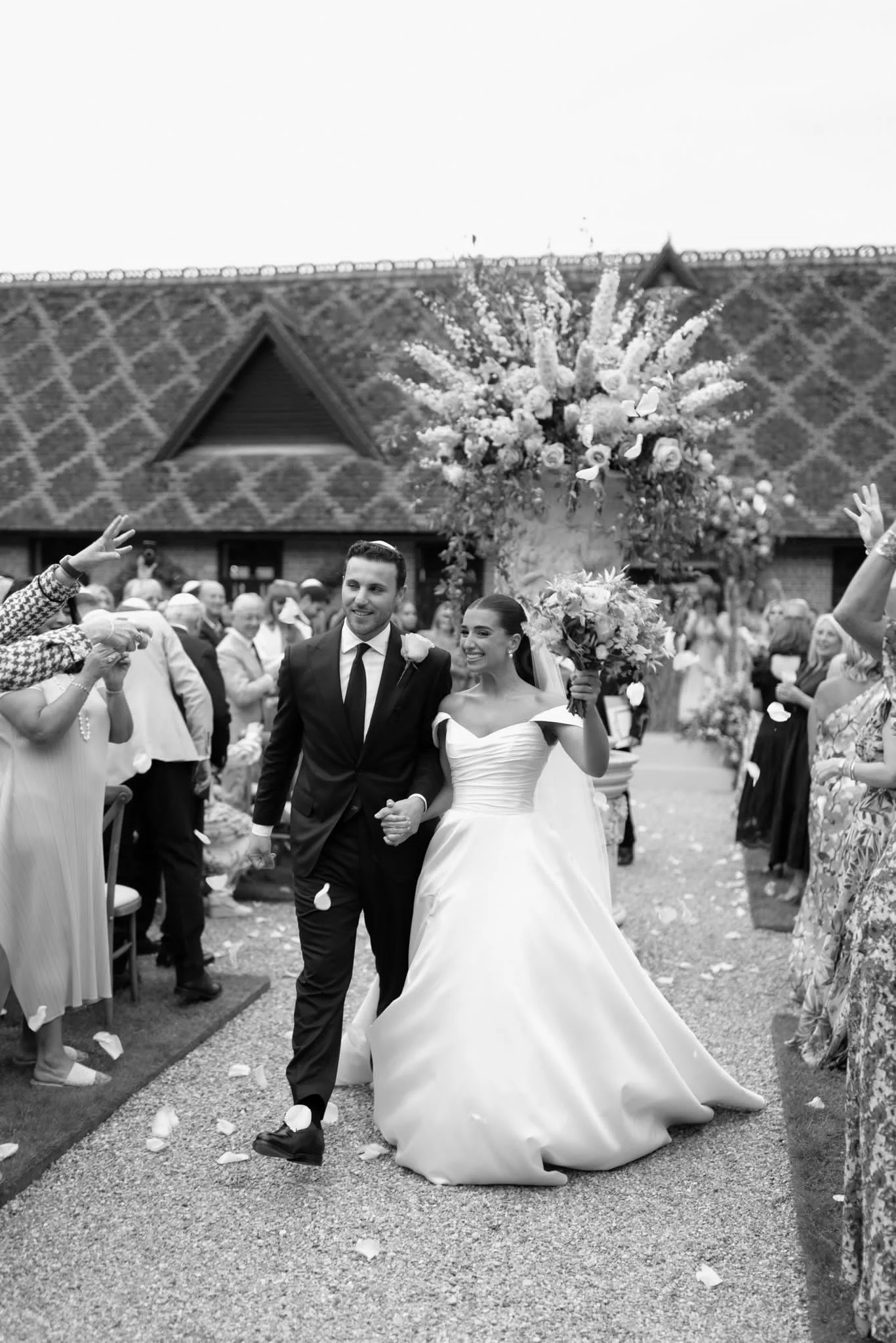 Black and white photo of a newlywed couple walking hand in hand. The bride is holding a large bouquet, and the groom is dressed in a suit. They are surrounded by guests, some reaching out, and an arch decorated with flowers and foliage is behind them