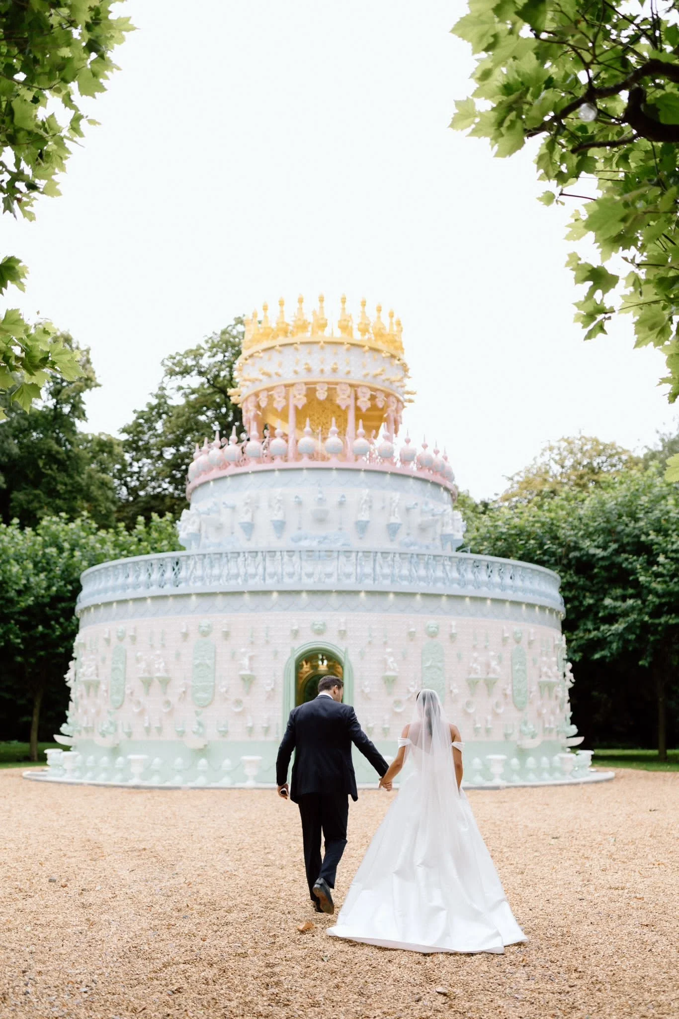 A bride and groom walking hand in hand towards a decorative pastel-colored church, surrounded by trees, during their wedding.