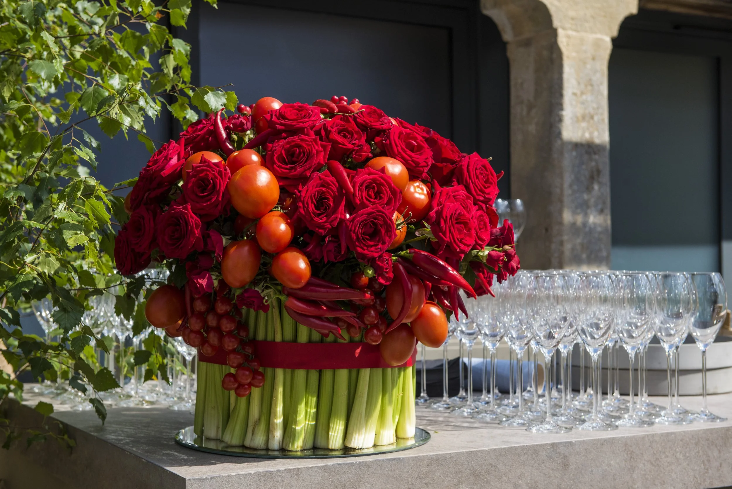 A large floral arrangement of red roses, orange tomatoes, and red chili peppers in a tall green and yellow vase, set on a counter with empty wine glasses beneath a stone archway.