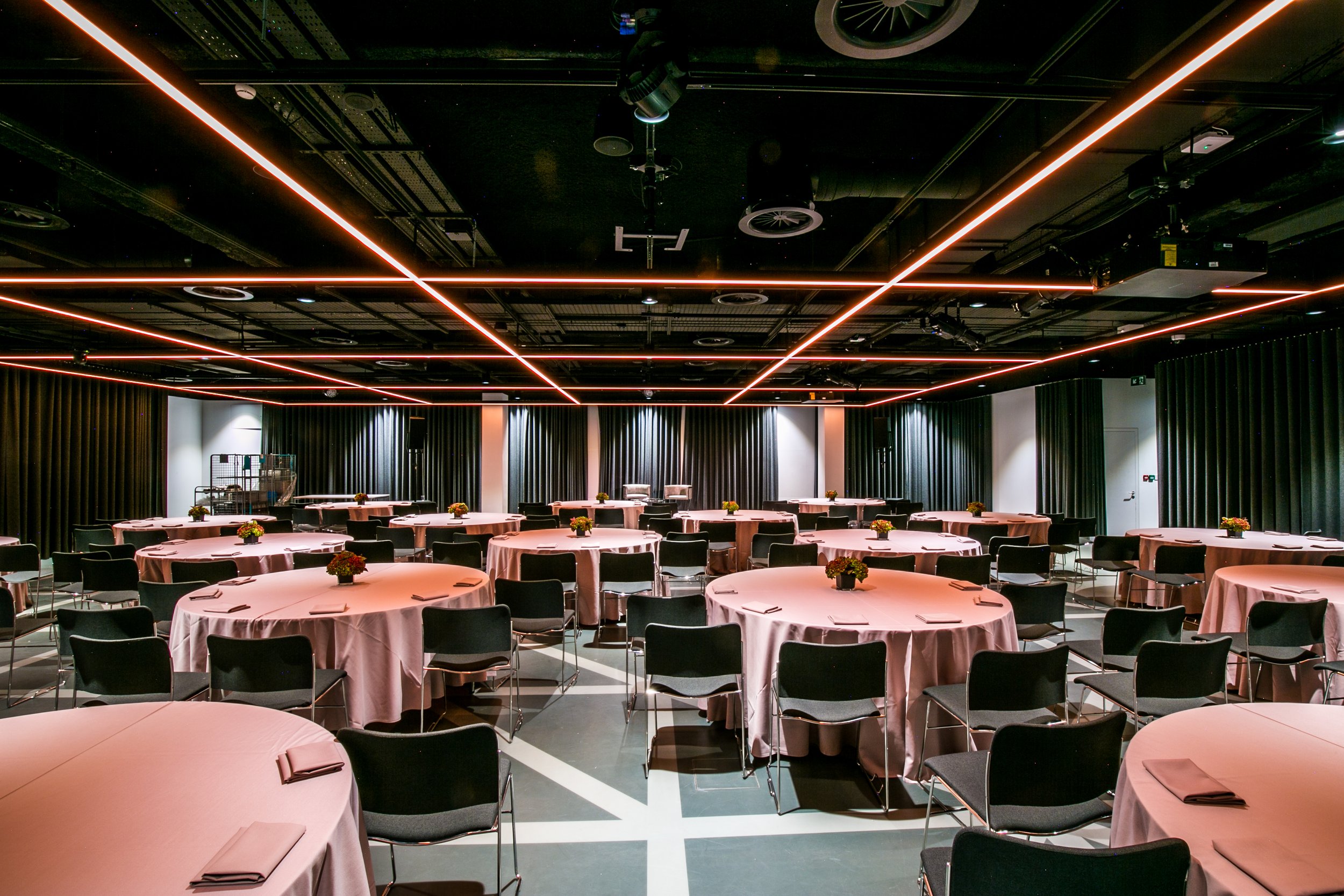 A conference or event room with round tables covered in pink tablecloths, each set with napkins and small floral centerpieces. Black chairs surround the tables. The ceiling features modern linear lighting and visible equipment such as speakers and ai