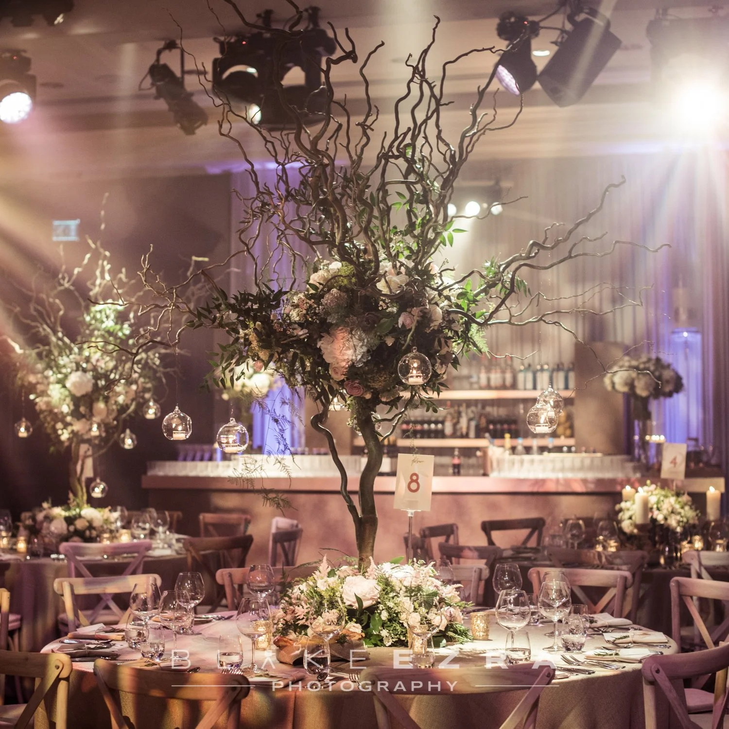Elegant event table with floral centerpiece featuring twisted branches and hanging glass candles, surrounded by wine glasses, plates, and candles in a warmly lit setting.