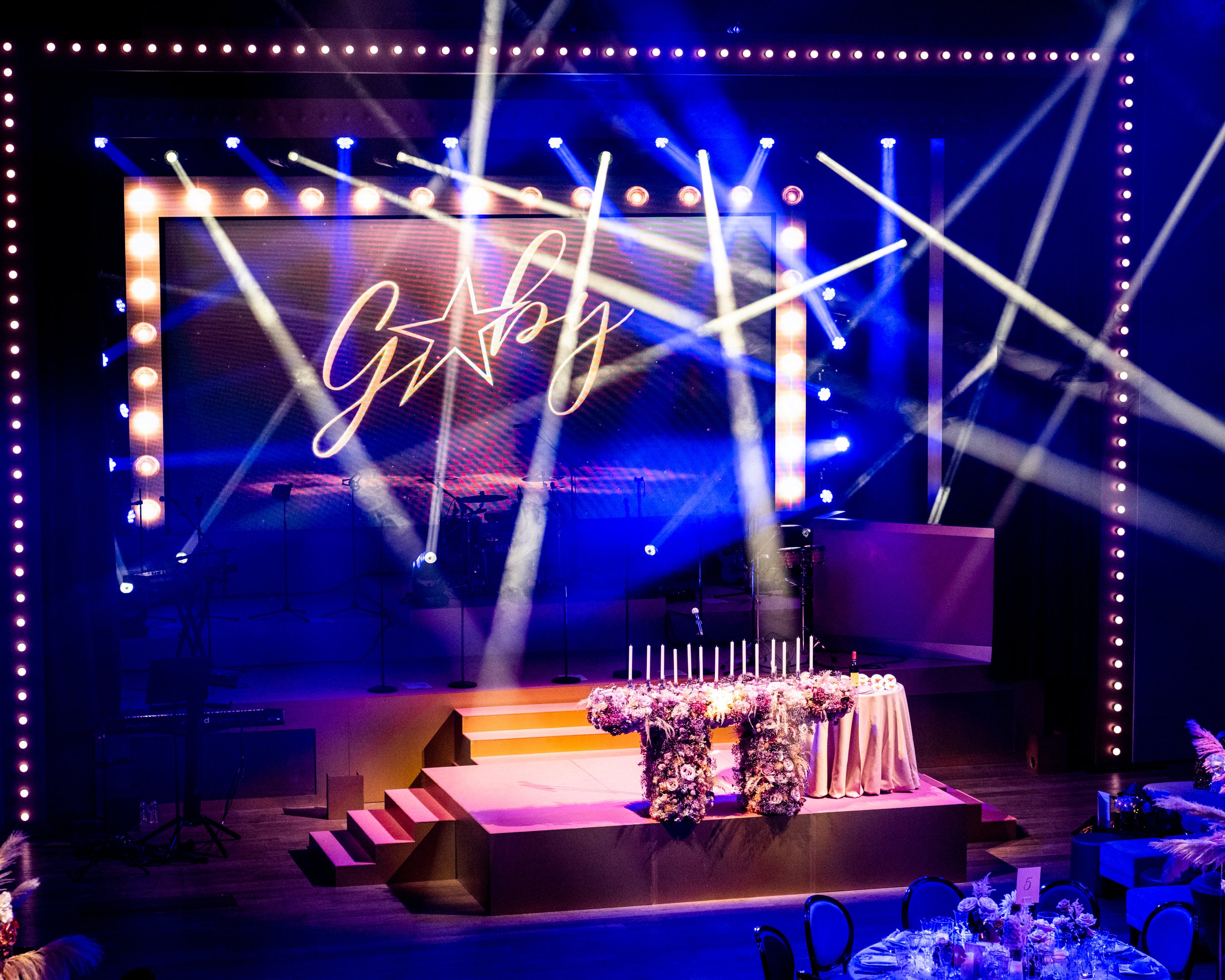 A decorated stage for a special celebration with flowers, a cake on a table, and a large screen displaying the message 'Nghi' with a star. The stage is illuminated with bright spotlights and surrounded by a border of lights. BAT MITZVAH AT BATTERSEA 