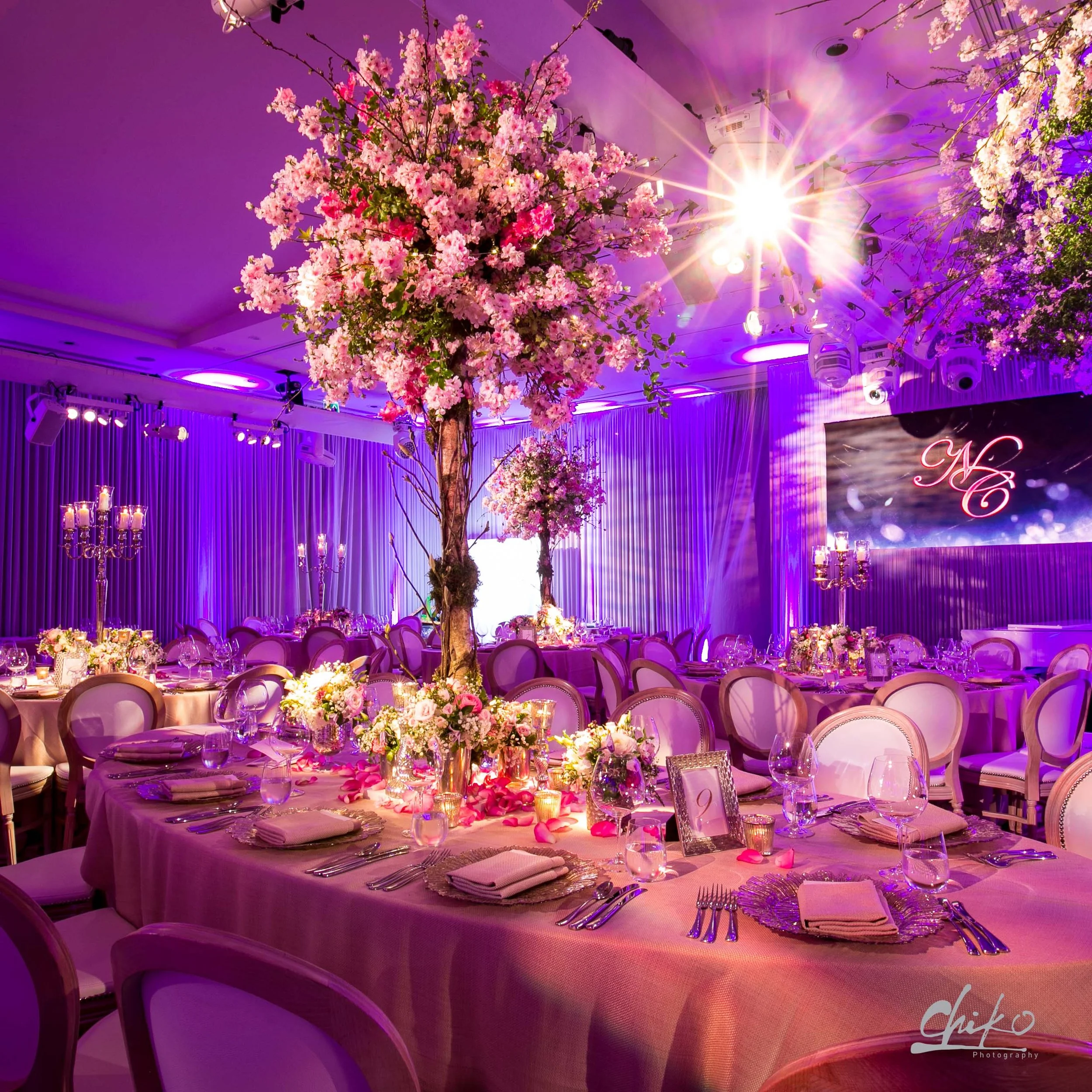 Elegant banquet hall decorated with pink cherry blossoms on trees, purple lighting, floral centerpieces, candles, and table settings for a formal event or wedding reception.