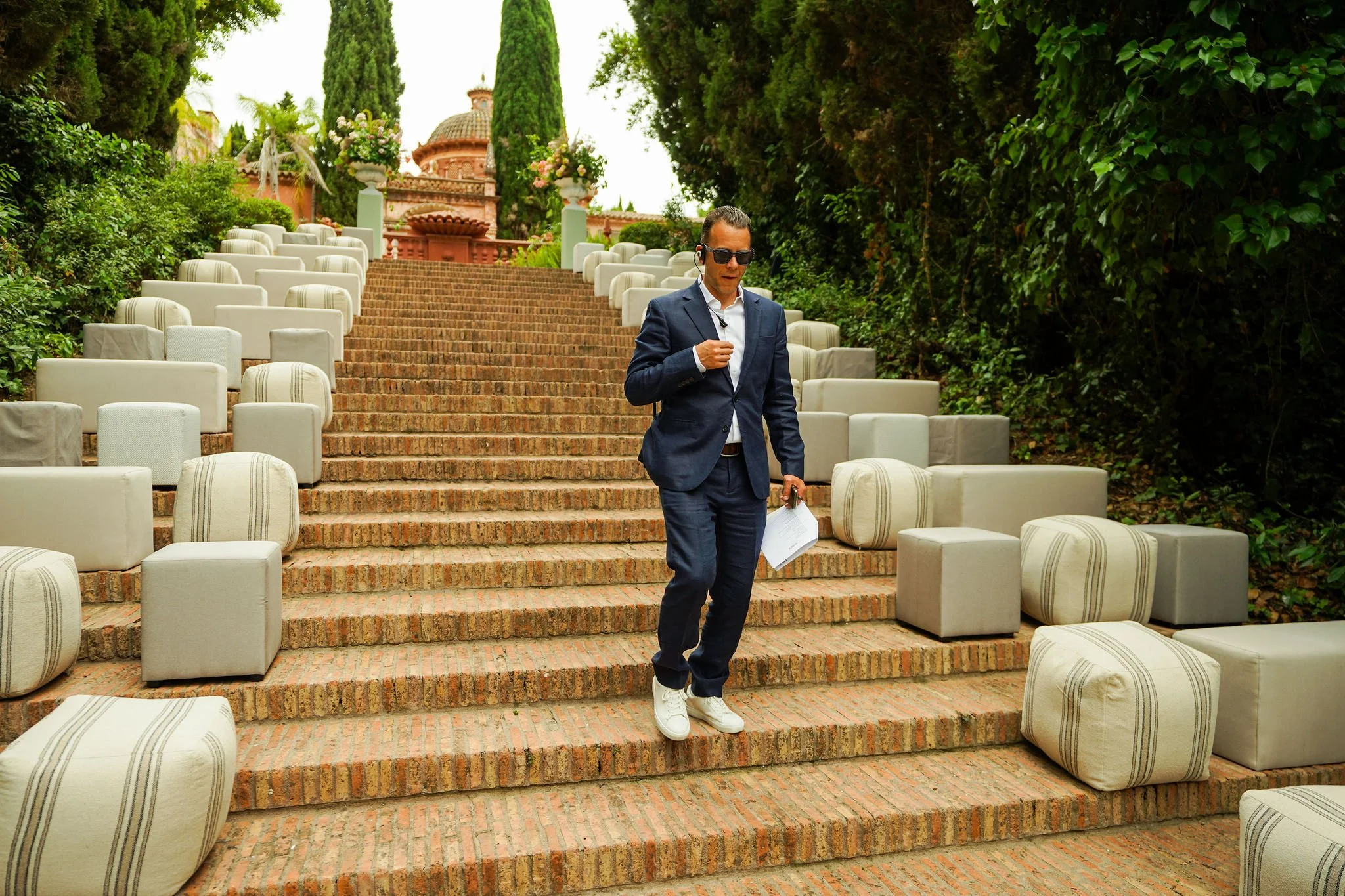 Laurie Weitzkorn director of JustSeventy  wearing sunglasses and headphones, walking down outdoor brick steps holding a paper and a gold-colored object, with rows of white padded chairs on both sides and lush green trees in the background.