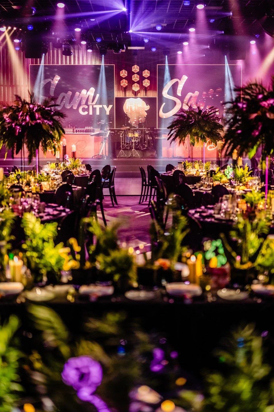 An event space with a stage, purple and pink lighting, and decorated tables with floral centerpieces. The stage features large screens with the words 'Dream City' and 'S' and a silhouette of a woman with jewelry. There are palm trees and other tropic