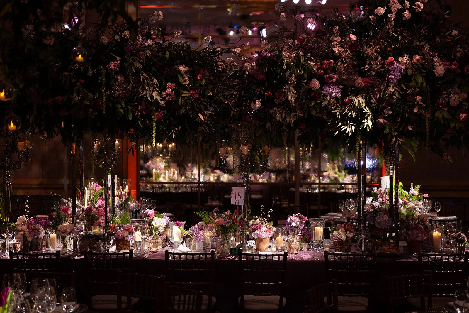An elegant event setup with tables decorated with pink and white flowers, candles, and glassware, under an elaborate floral canopy with dim lighting at The Peninsula London