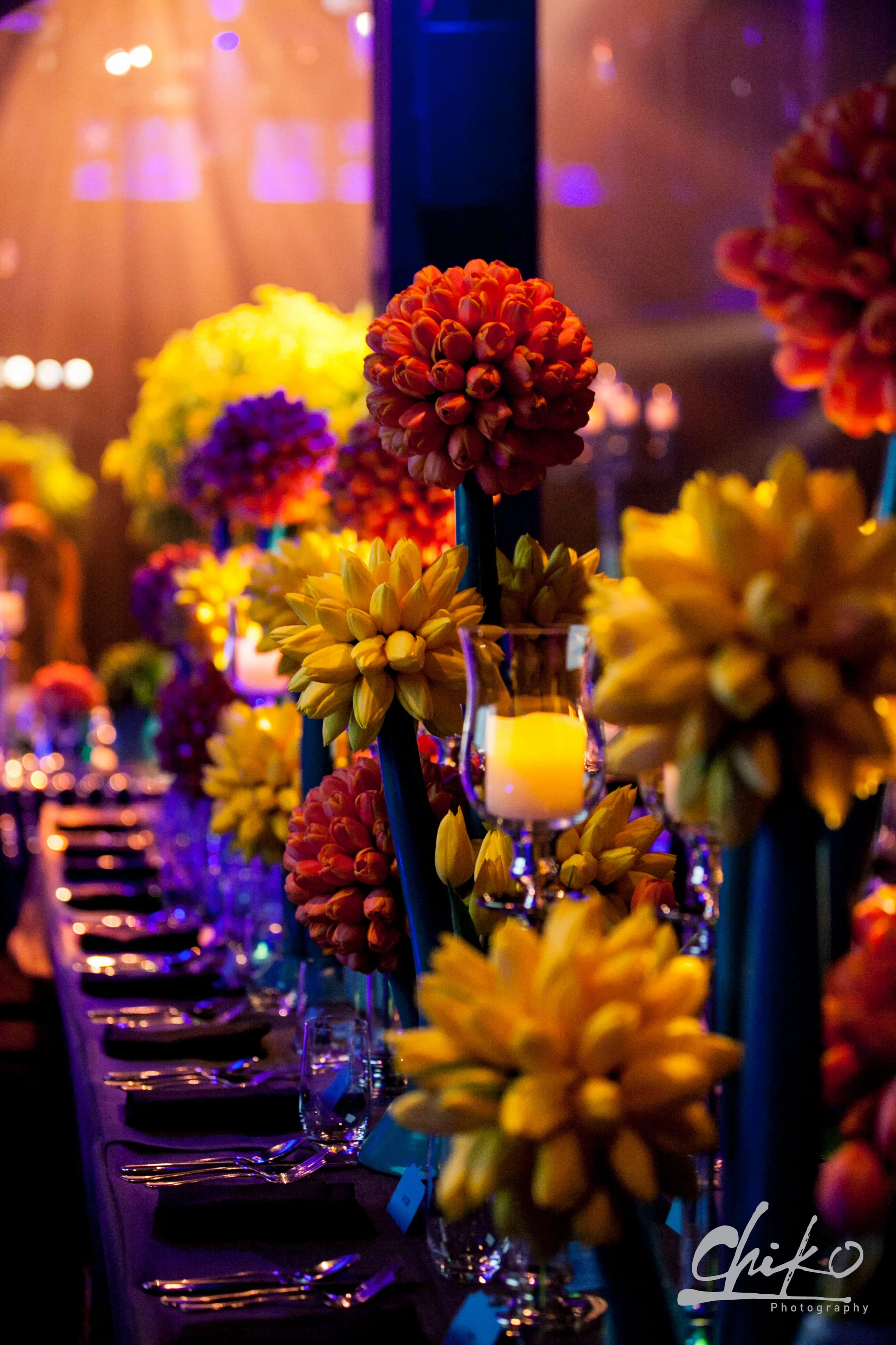 A dimly lit banquet table decorated with vibrant orange, purple, and yellow flowers and floating candles in glass holders, arranged for a special event or celebration.
