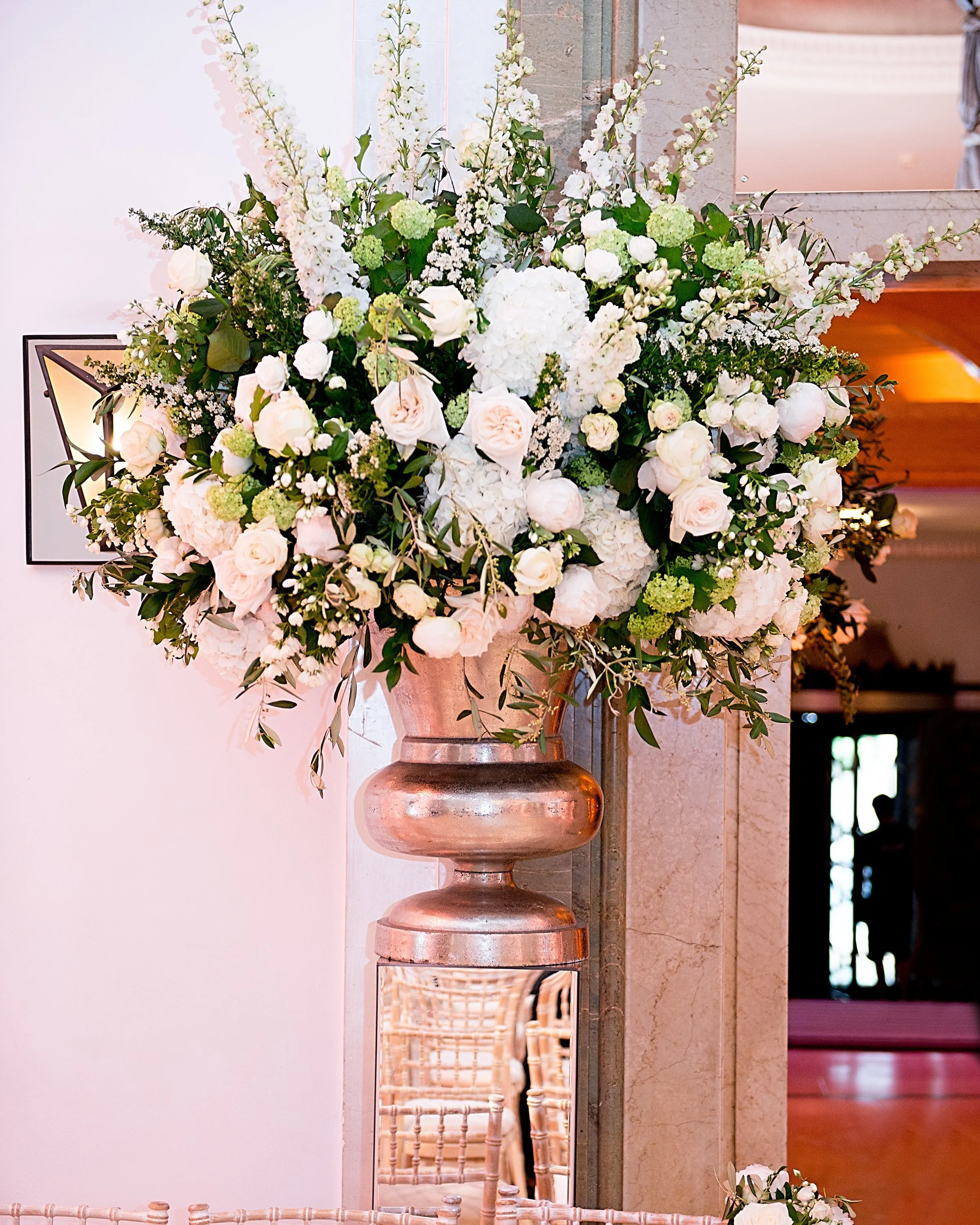 A large floral arrangement with white roses, hydrangeas, and other white flowers and green foliage in a copper urn, positioned next to a mirror on a beige marble surface, inside a building.